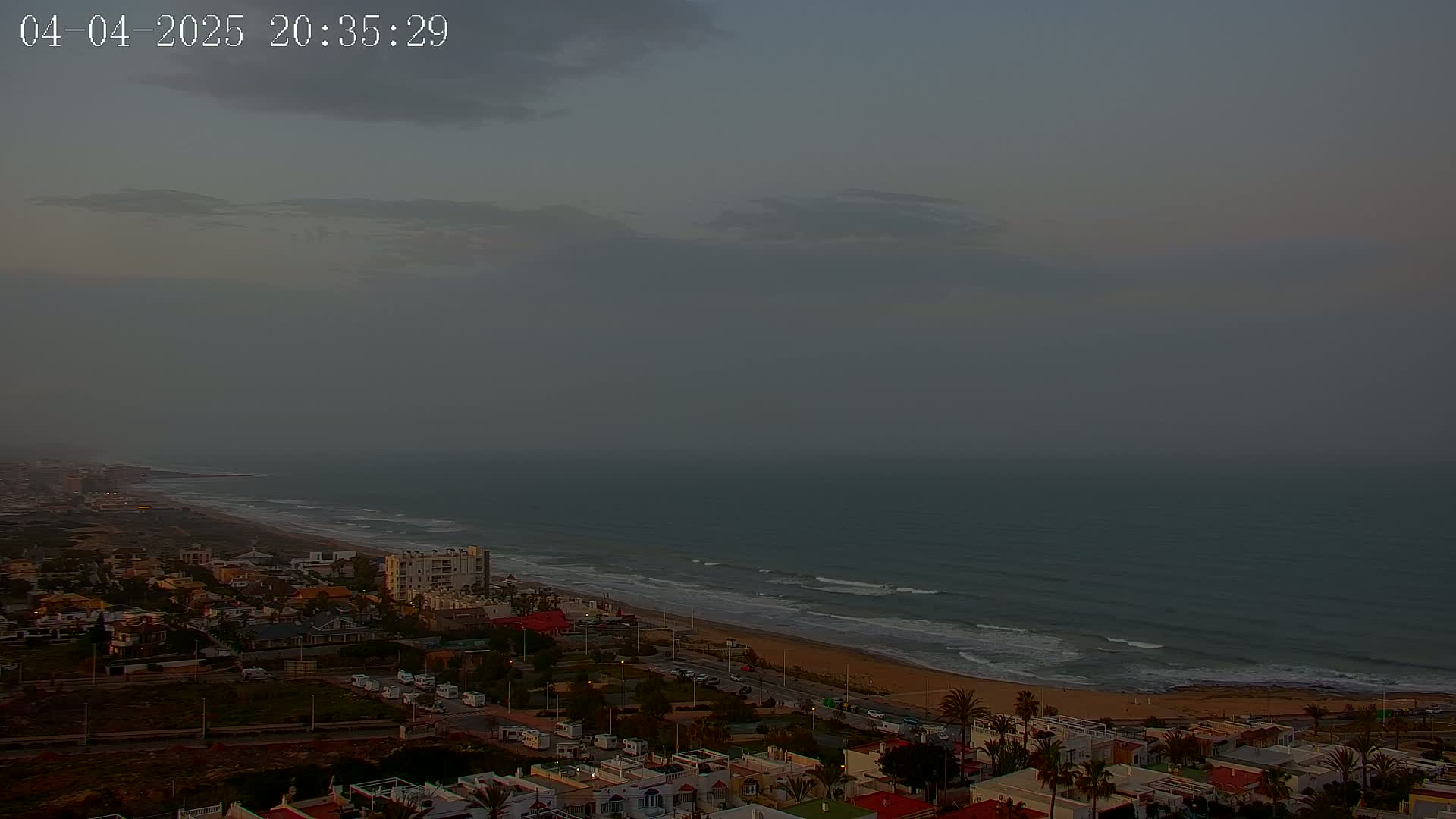 An overcast sky hangs over a coastal town with a sandy beach and gently rolling waves at dusk.
