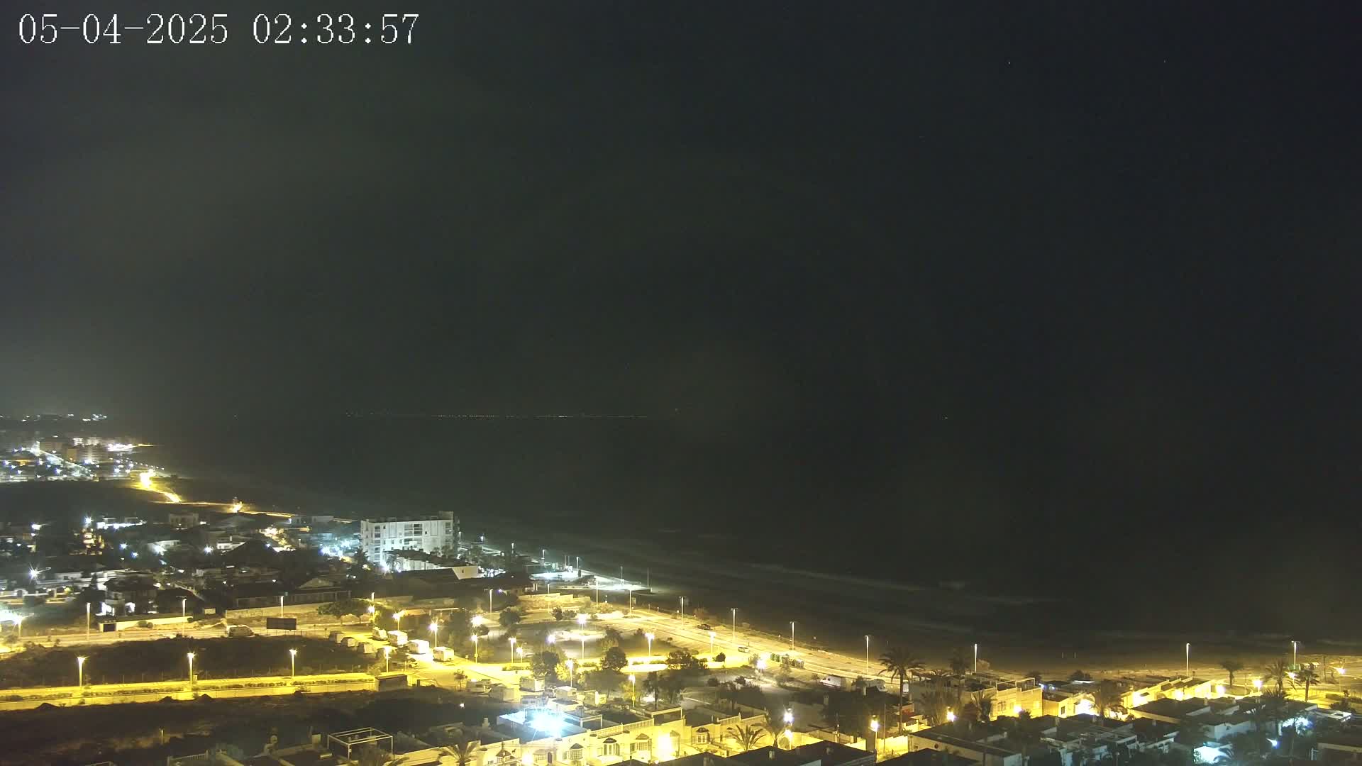 A nighttime coastal town with illuminated streets and buildings overlooks a dark ocean with gently lapping waves under a clear, starlit sky.