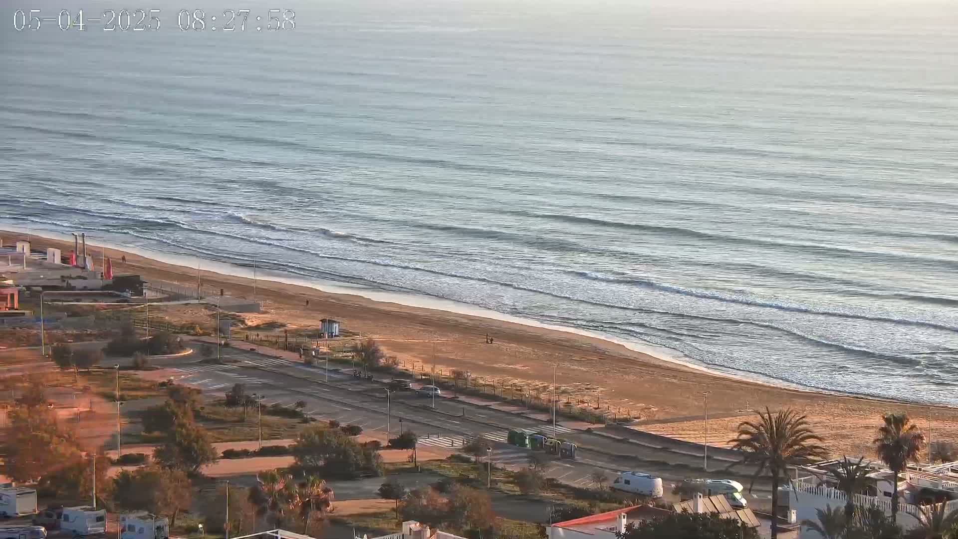 An aerial view shows a sandy beach with gently rolling waves on a calm, clear day, adjacent to a coastal road lined with vegetation and parking.