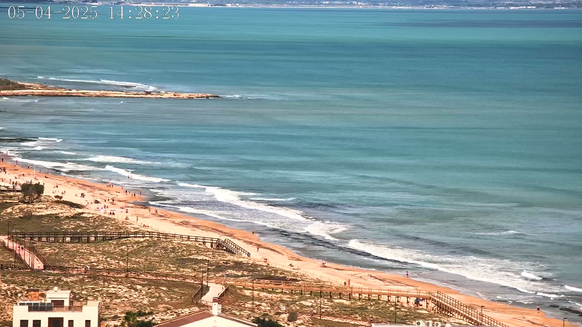 A sandy beach with people sunbathing and walking along a boardwalk is bordered by a calm, teal ocean with small waves breaking on the shore.