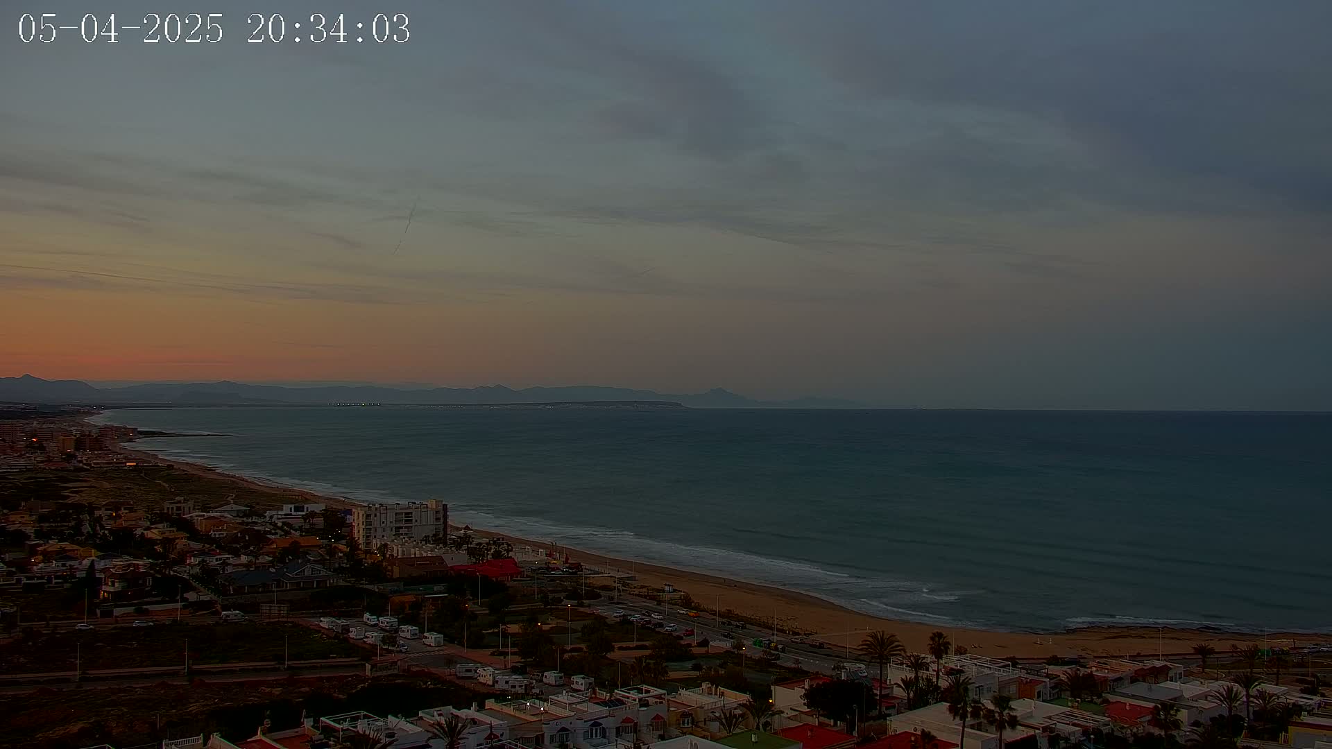A coastal town at sunset overlooks a calm ocean with a distant mountain range under a cloudy sky.