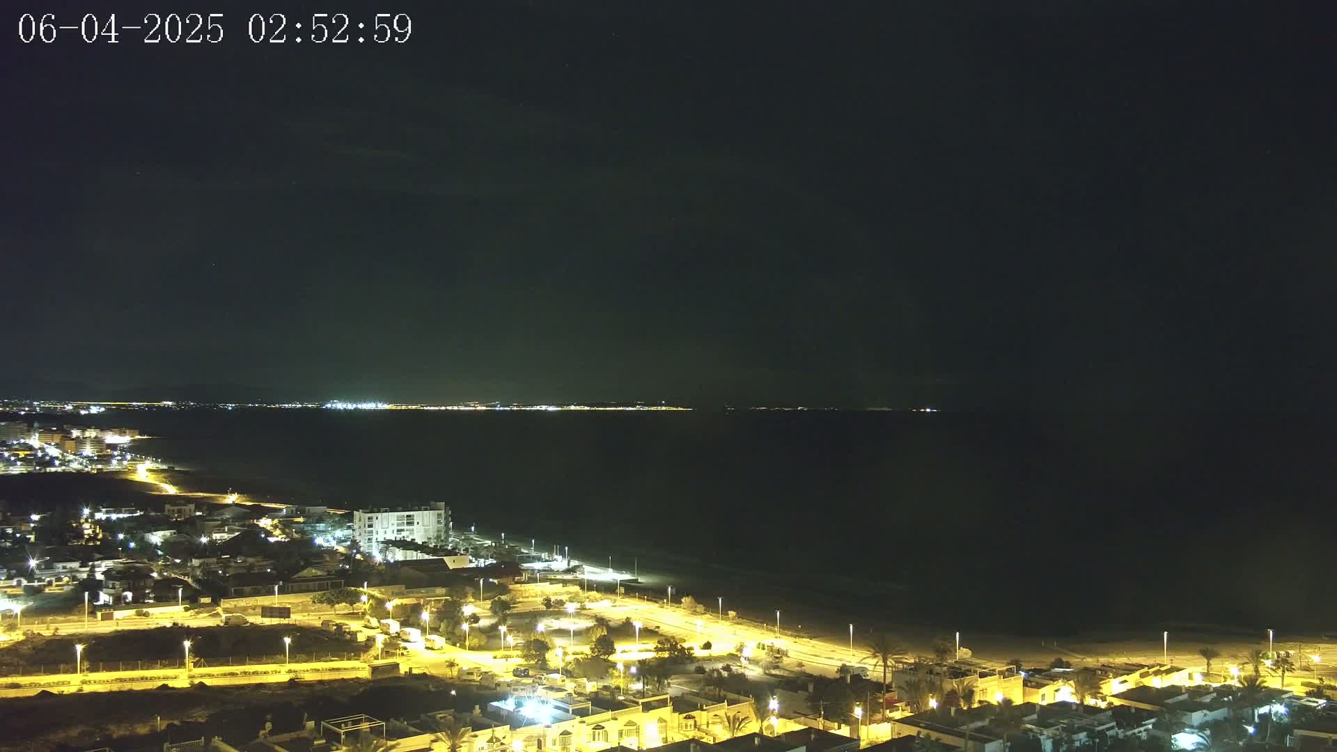 A nighttime coastal scene shows a dark ocean with faintly visible lights on the far shore, a brightly lit beach, and a residential area illuminated by streetlights.