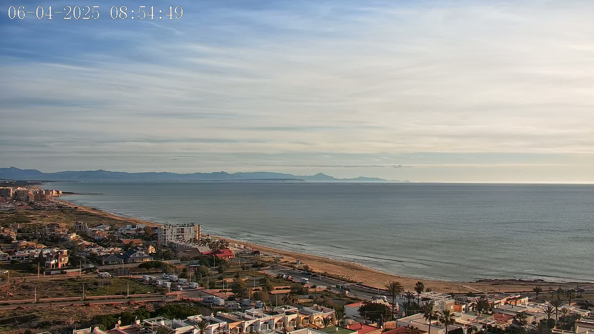 A coastal town with a sandy beach and calm sea stretches towards distant mountains under a mostly clear, light blue sky.