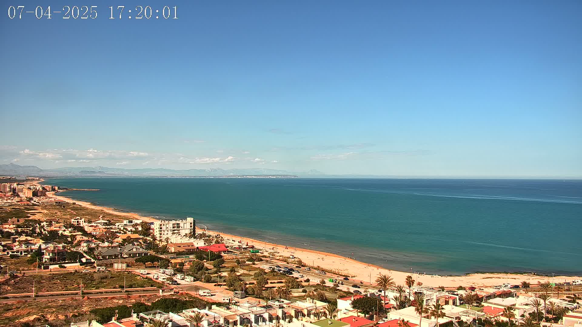 A coastal town with sandy beach and teal ocean stretches to distant mountains under a clear blue sky.