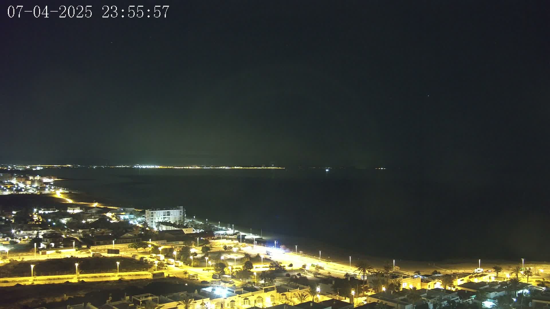 A nighttime coastal scene shows a dark sea under a clear, dark sky, with a town's illuminated buildings and streets along the shoreline.