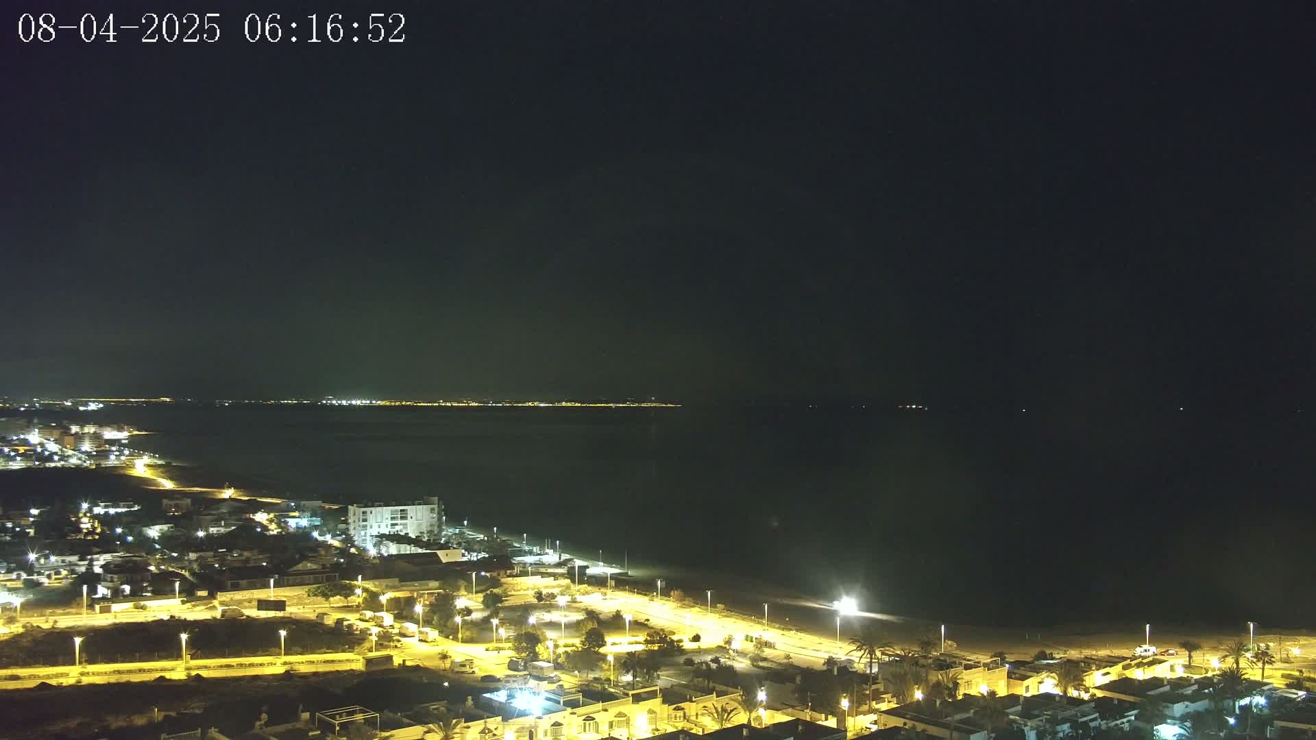 A nighttime coastal view shows a dark sea, a dimly lit beach, and a town with illuminated streets and buildings under a clear, dark sky.