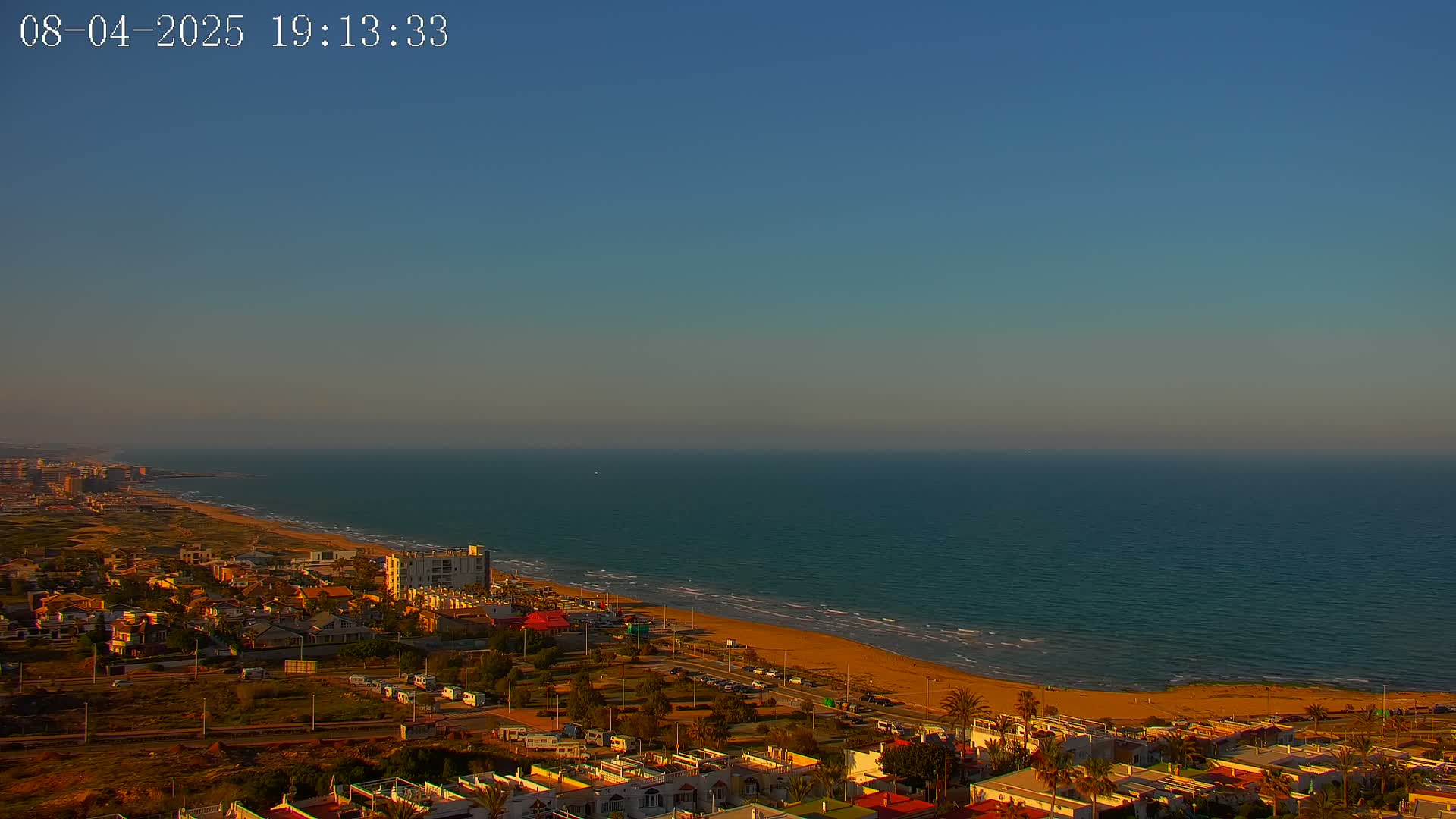 An aerial view shows a sandy beach meeting a calm ocean under a clear, twilight sky, with a residential area situated behind the beach.