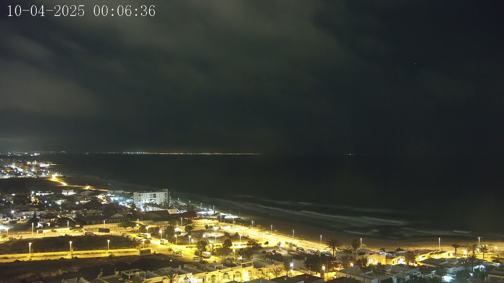 A nighttime coastal town is illuminated, with a dark, cloudy sky above and gentle waves lapping a sandy beach.