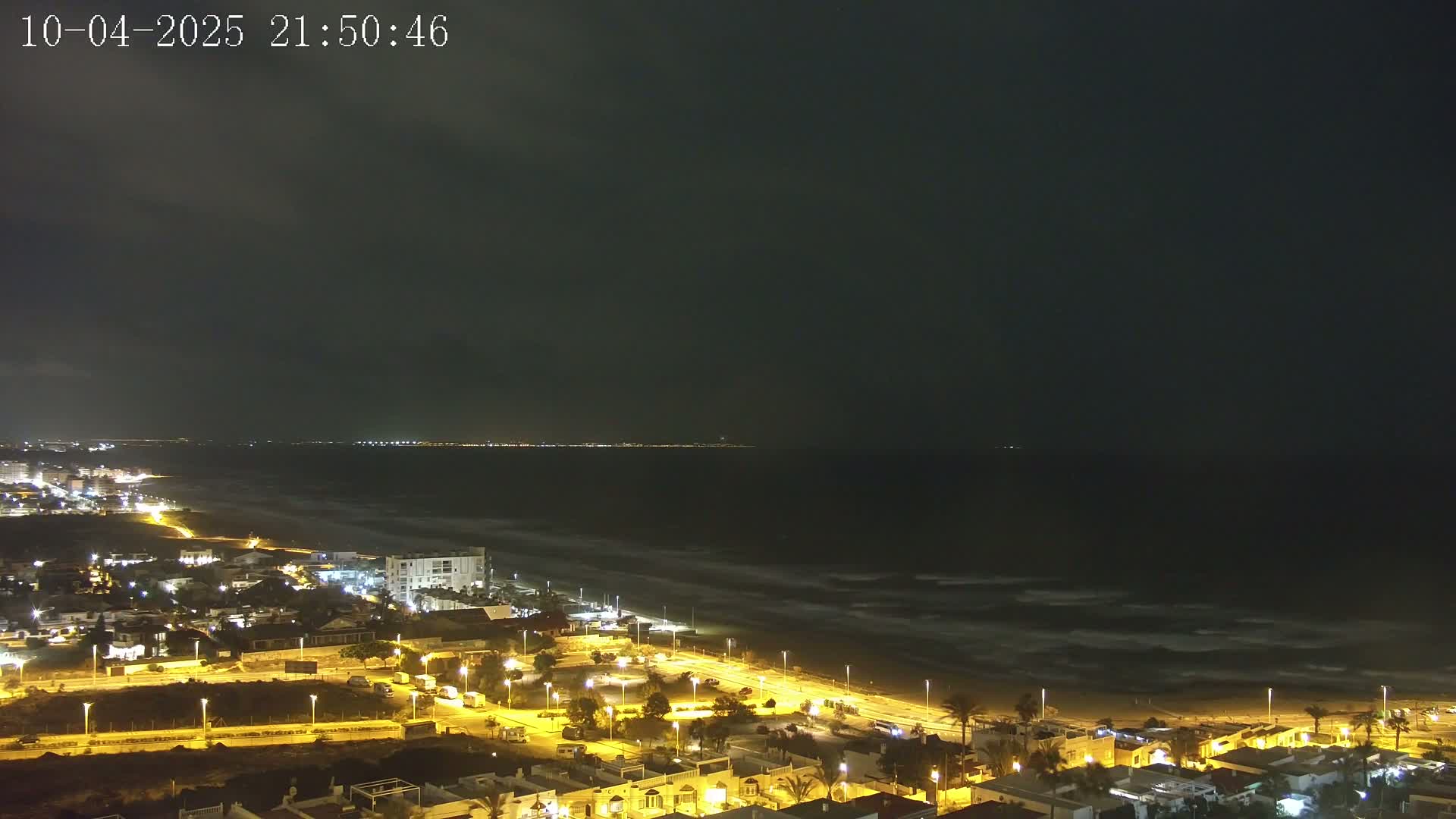 A nighttime coastal town brightly lit, overlooking a dark ocean with small waves under a mostly cloudy sky.