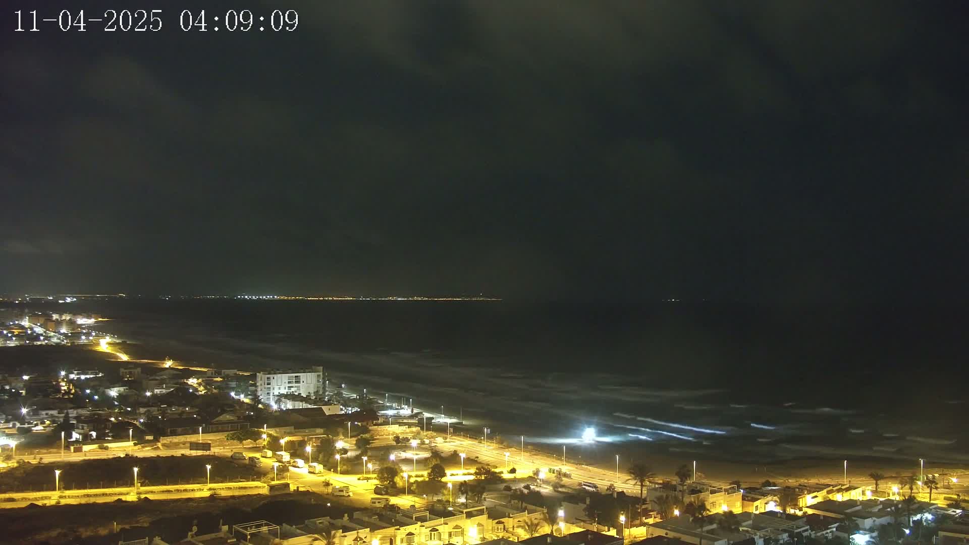 A nighttime coastal town is illuminated, with moderately wavy water visible under a dark, cloudy sky.