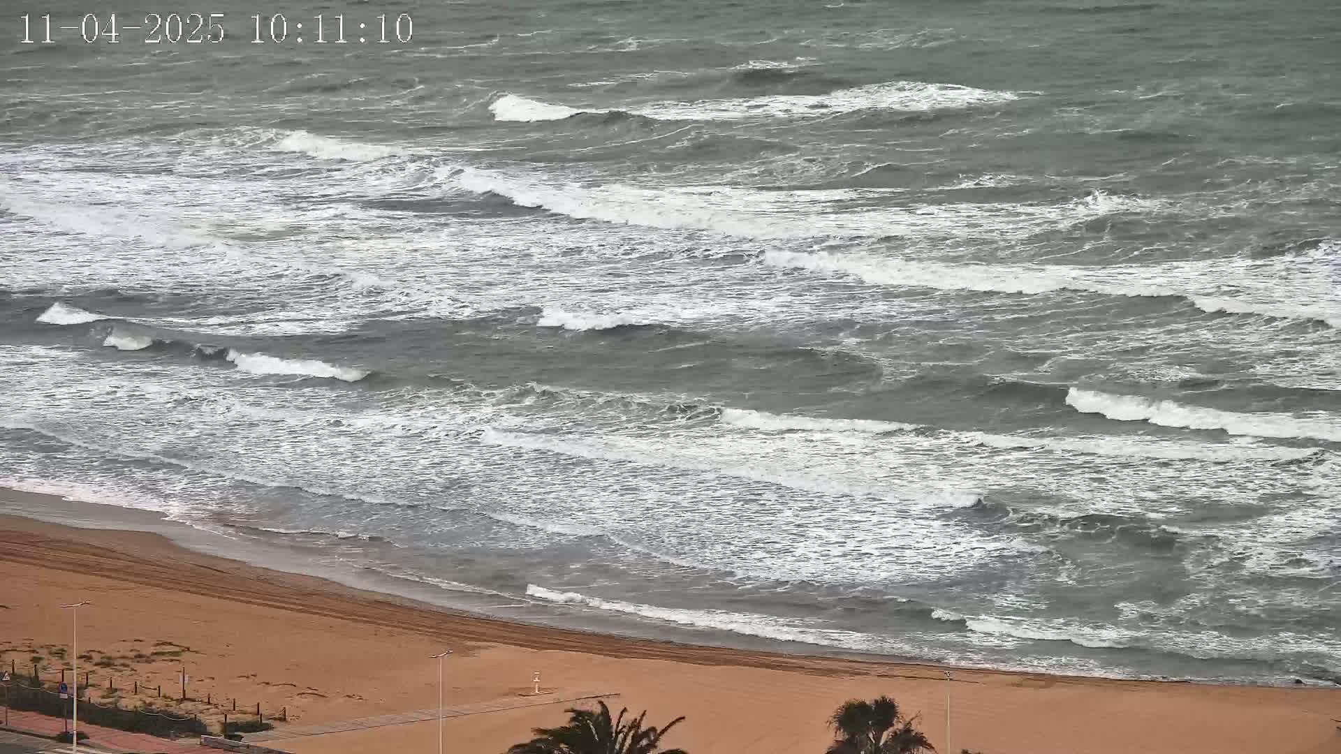 A sandy beach meets a gray, choppy sea with moderate waves under an overcast sky.