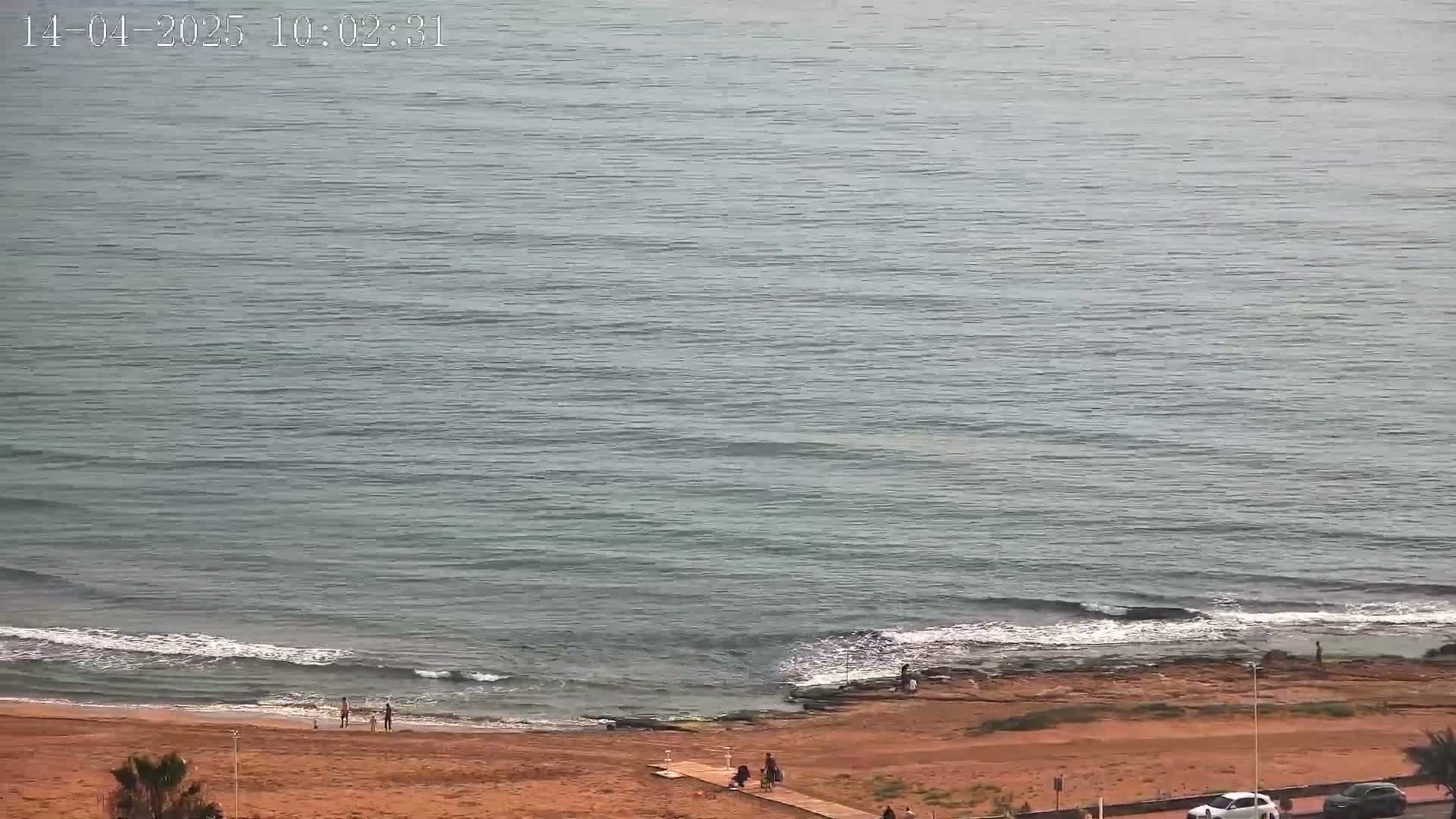 A mostly calm sea with small waves laps a sandy beach where a few people are scattered.