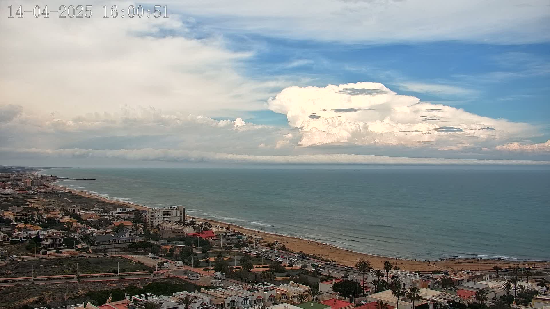 A coastal town overlooks a wide sandy beach and calm ocean under a partly cloudy sky with large cumulus clouds.