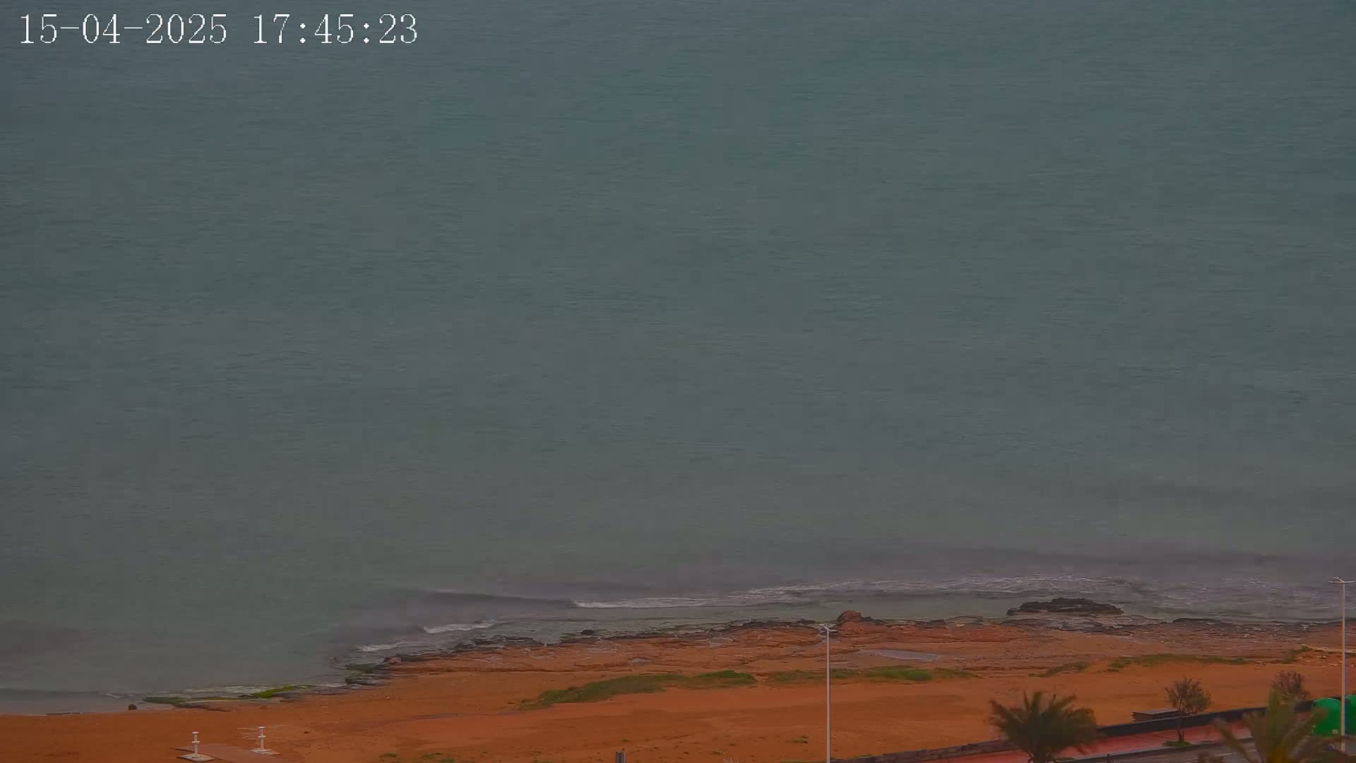 A mostly overcast sky overlooks a sandy beach and calm, gray-green ocean with small waves breaking on the shore.
