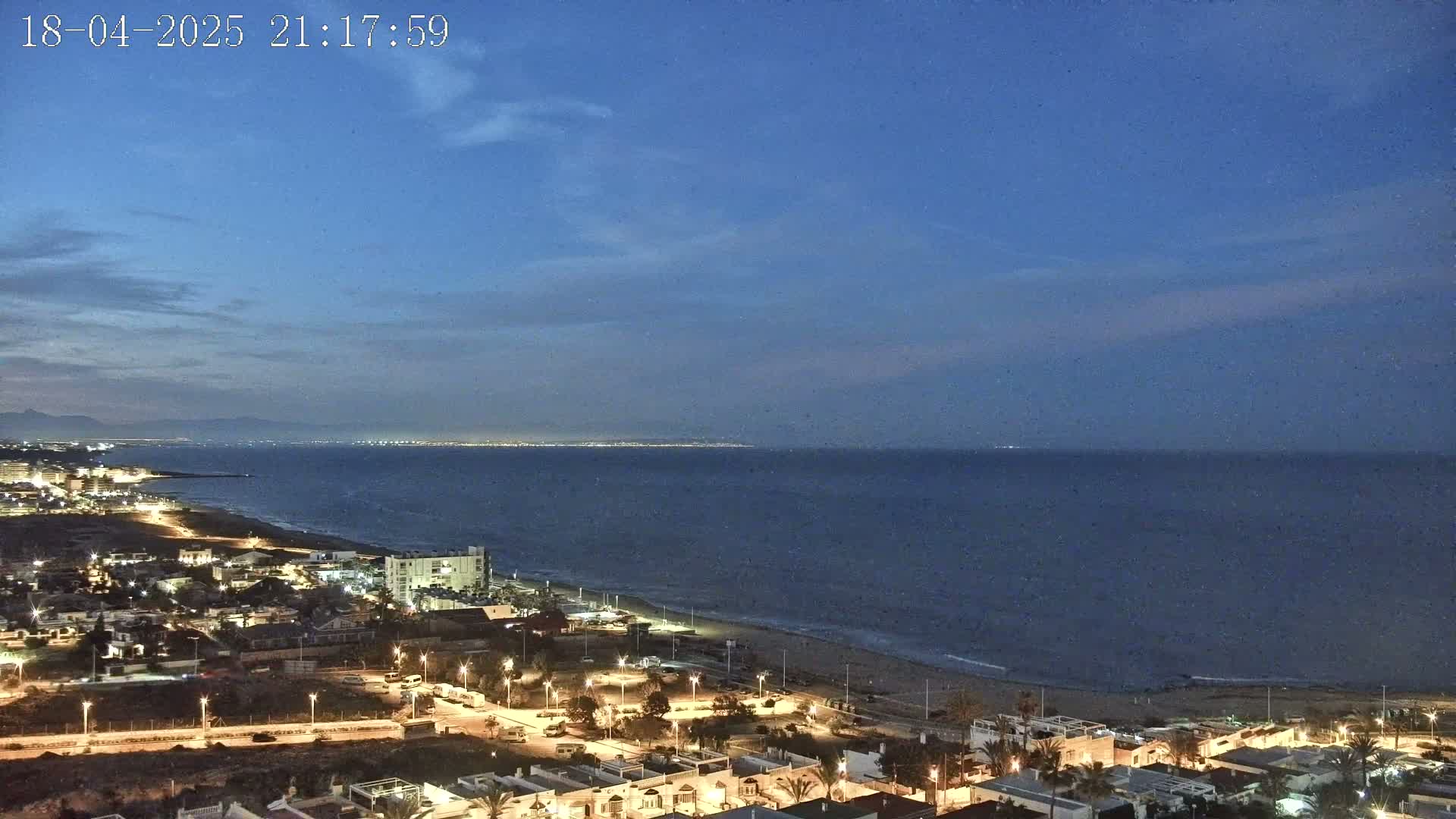 A coastal town at twilight is illuminated at night, overlooking a calm sea under a mostly clear, dark blue sky.