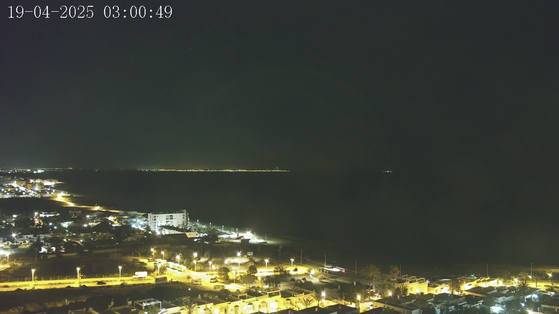 A nighttime coastal scene shows a dark ocean with a faintly lit distant shoreline, and a closer, brightly lit residential area with streetlights illuminating houses and roads.