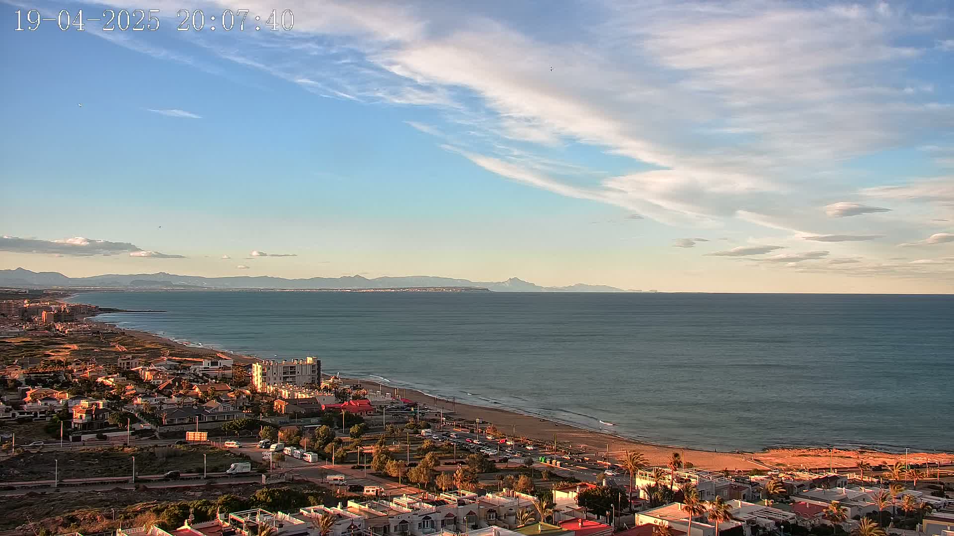 An expansive coastal view shows a sandy beach, a town, and a calm ocean under a mostly clear sky with scattered clouds at sunset.