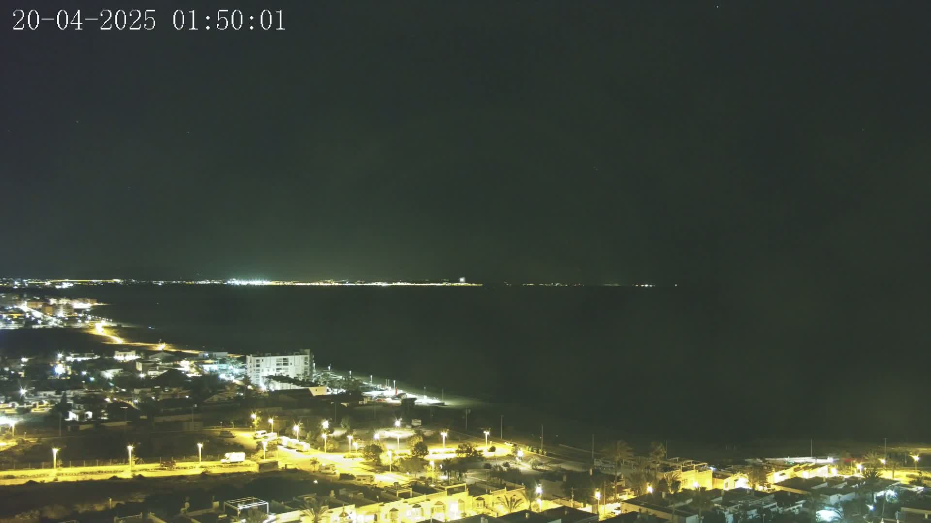 A nighttime coastal town is illuminated, overlooking a dark body of water with a distant, faintly lit shoreline under a clear, starlit sky.