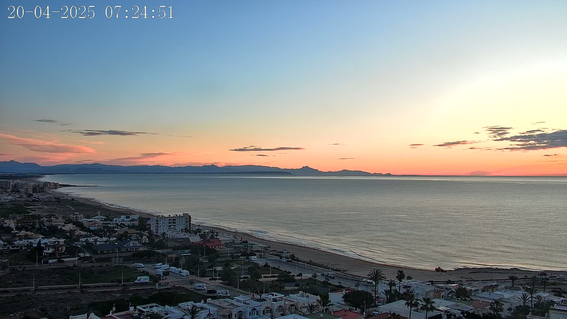 A coastal town at sunrise, with a calm sea extending to distant mountains under a pastel sky.