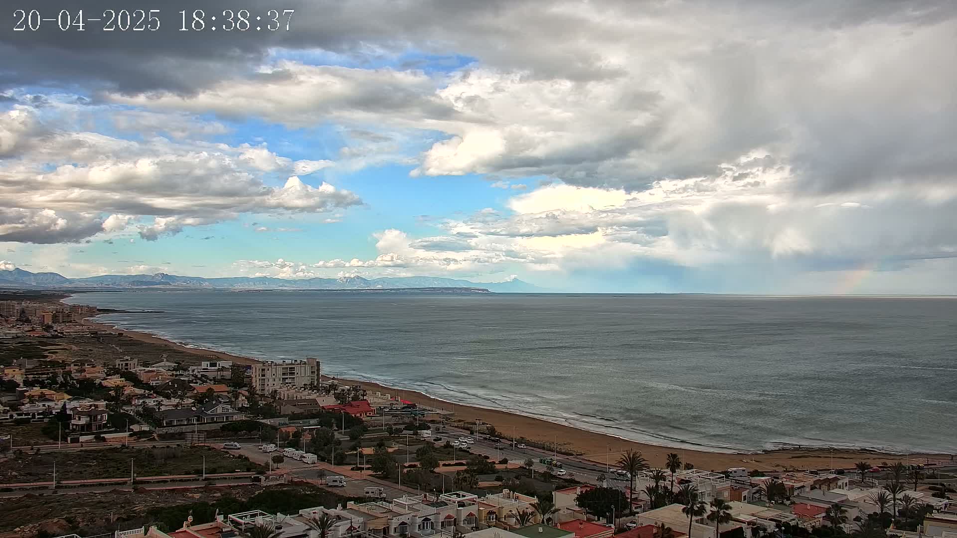 A coastal town overlooks a wide, grey ocean under a partly cloudy sky with a faint rainbow on the horizon and distant mountains.