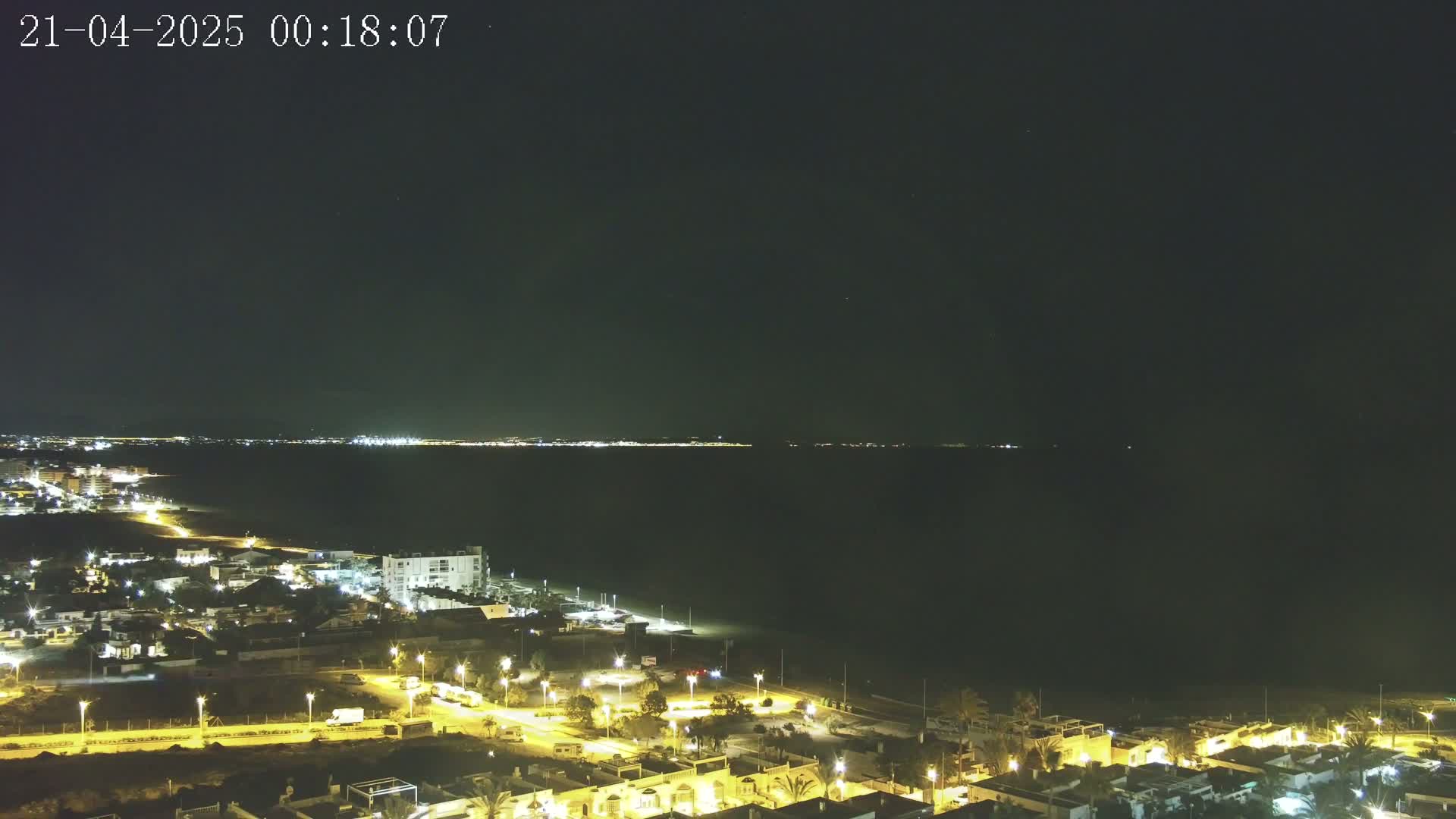 A nighttime coastal town is illuminated, overlooking a dark body of water with a distant city skyline faintly visible under a clear, starry sky.