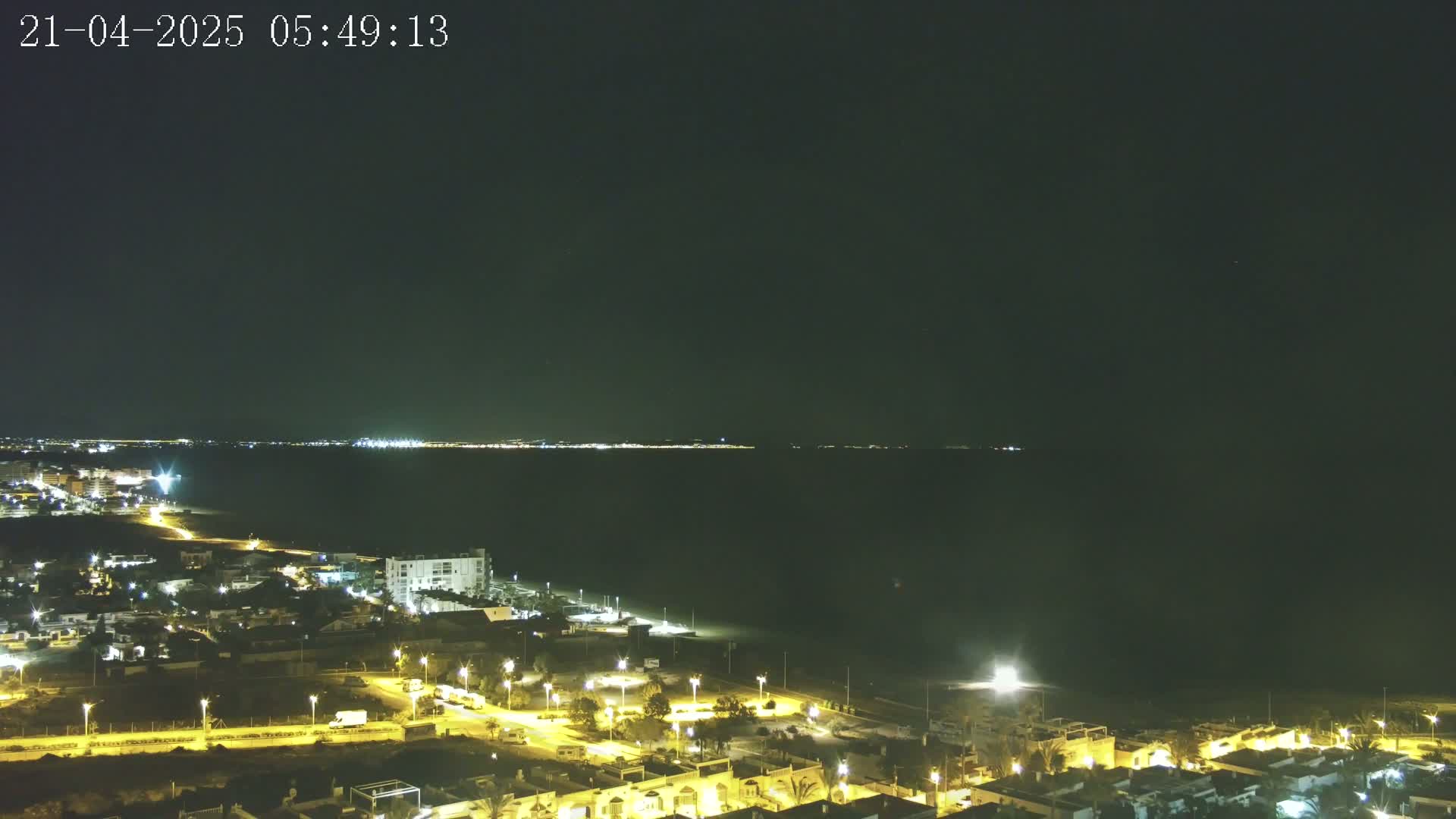 A nighttime coastal scene shows a dark body of water with a distant, faintly lit cityscape on the horizon, and a closer, brightly lit residential area with streets and buildings along the shoreline.