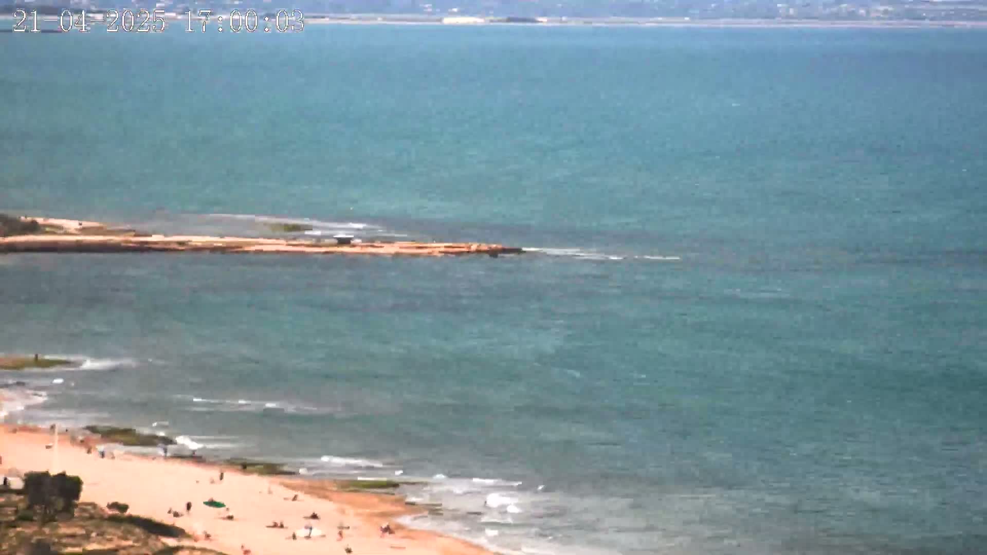 A sandy beach with a few people on it slopes down to a calm, teal sea extending to a distant shoreline under a clear sky.