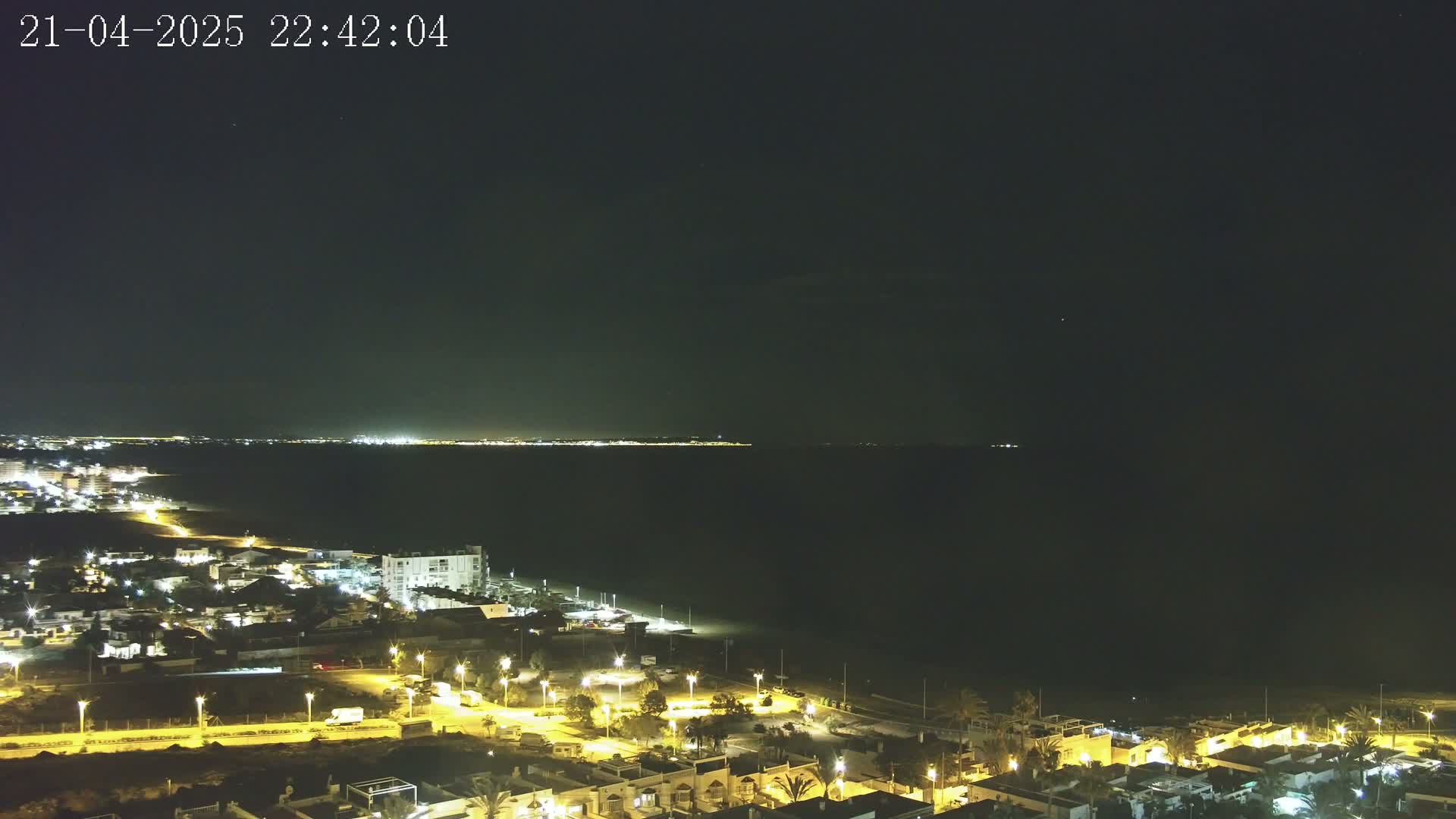 A nighttime coastal scene shows a dark body of water with a faintly lit distant shore, and a closer, illuminated town with streetlights bordering the beach.