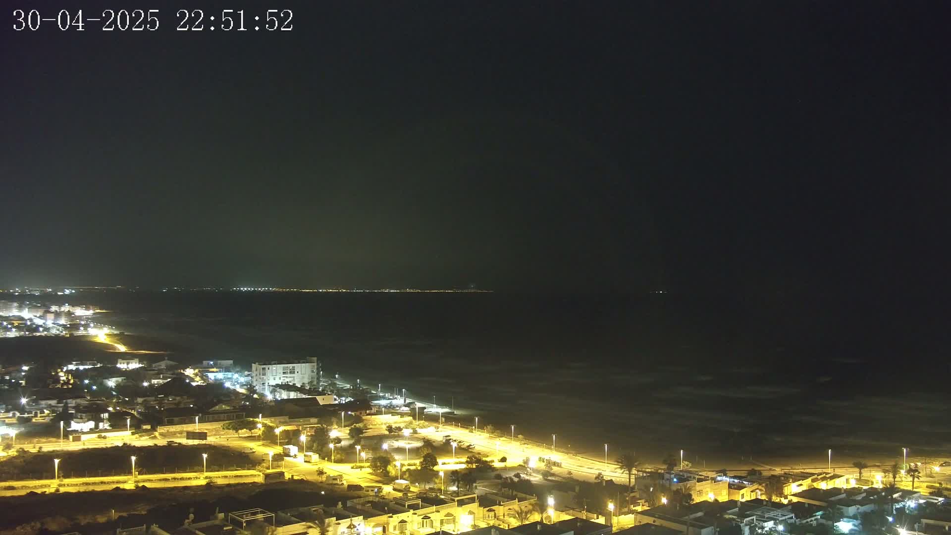 A coastal town at night is illuminated, with a dark, wavy ocean extending to a dimly lit horizon under a clear, dark sky.