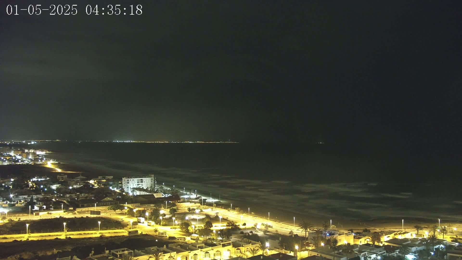 A nighttime coastal town with illuminated streets and buildings overlooks a dark ocean with gentle waves under a mostly clear sky.