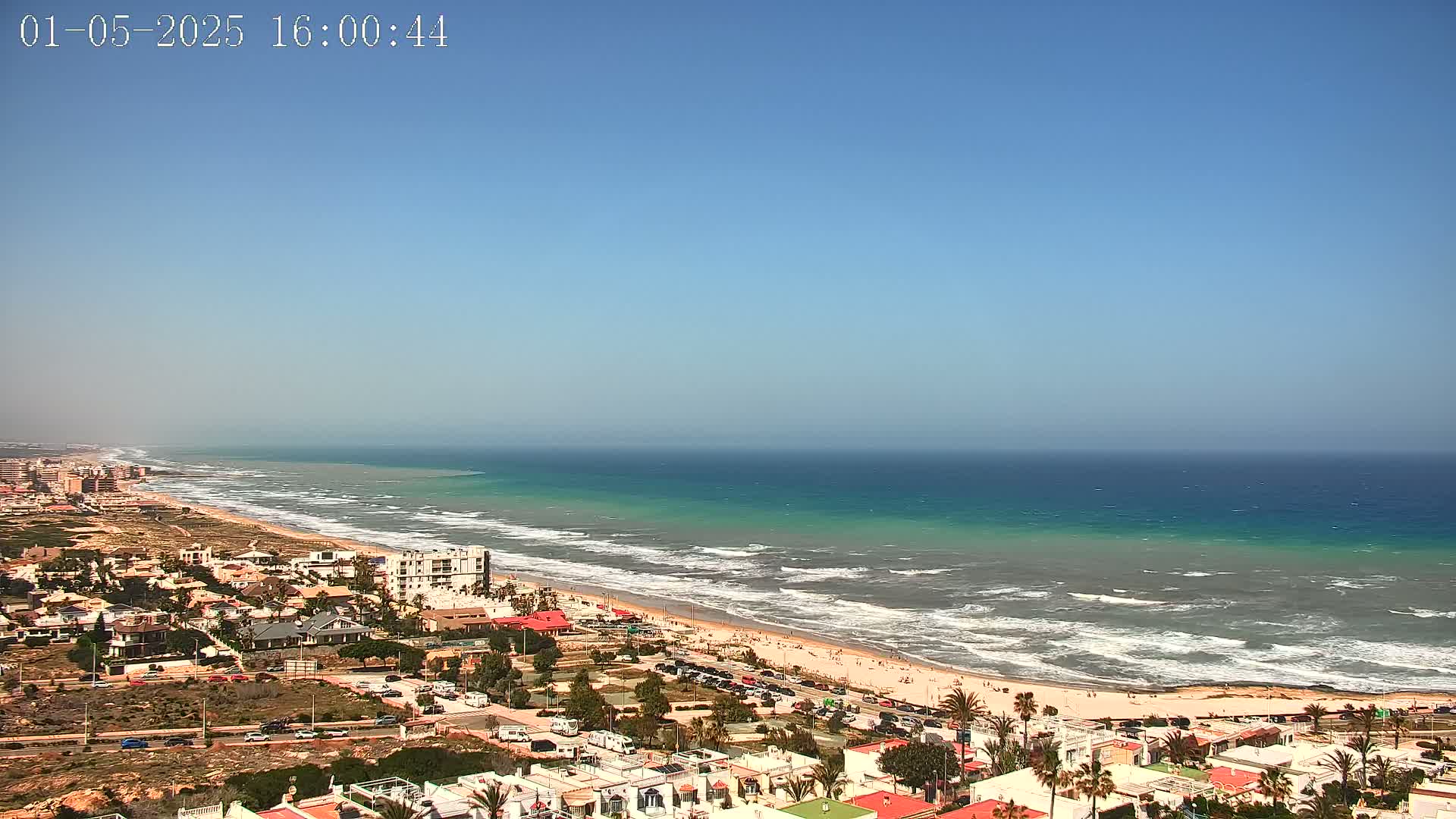 A coastal town overlooks a long sandy beach with moderate waves under a clear blue sky.