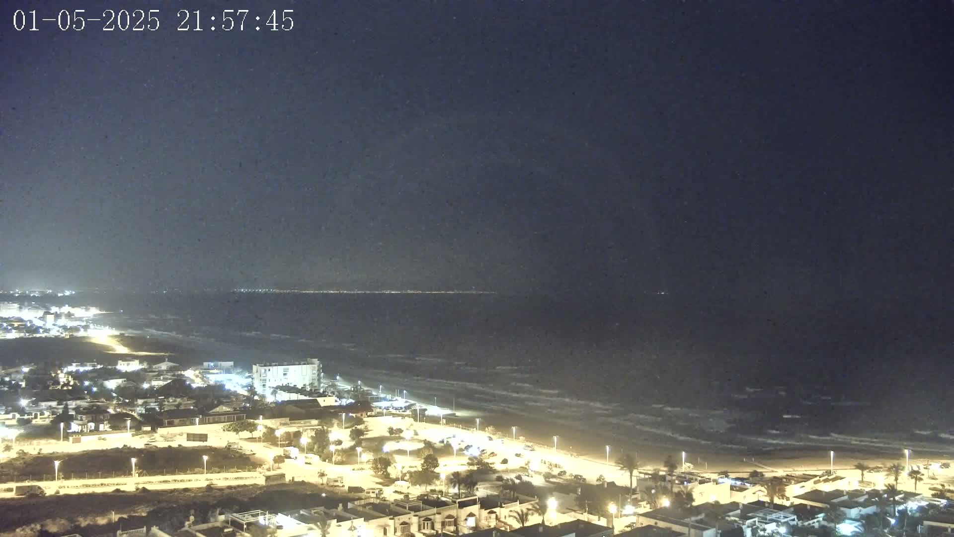 A nighttime coastal scene shows a dark ocean with waves under a clear sky, overlooking a beach and a town with illuminated buildings.