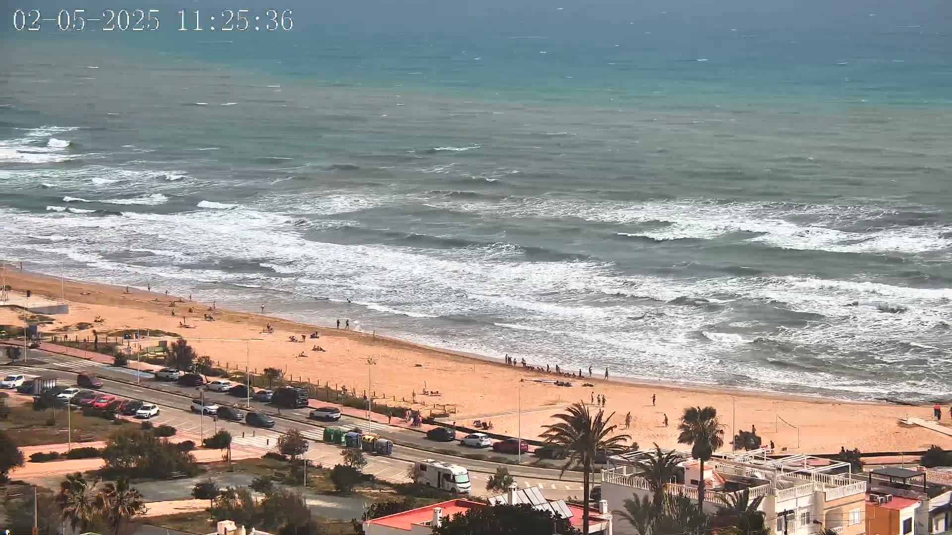 A sandy beach with scattered people is bordered by a road lined with parked cars and buildings, all overlooking a slightly choppy sea under a partly cloudy sky.