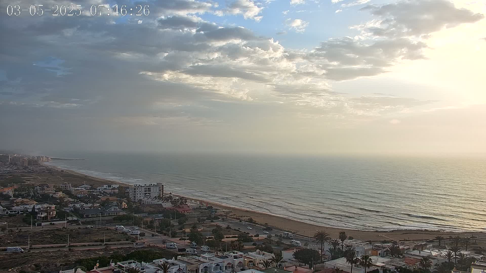 A hazy sunrise illuminates a mostly cloudy sky over a calm ocean, beach, and coastal town.