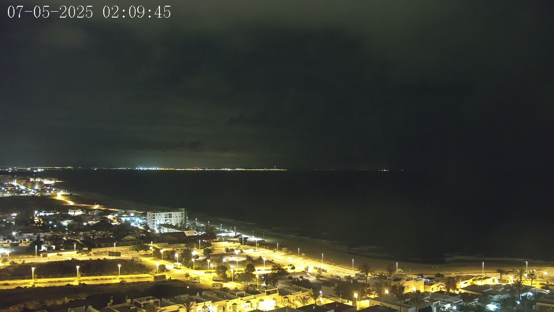 A nighttime coastal town is illuminated, with a dark, cloudy sky above and a calm, dark sea extending to a distant, faintly lit shoreline.
