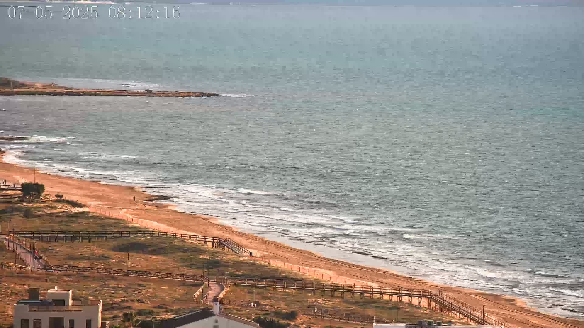 A sandy beach with gentle waves and wooden walkways is seen from an elevated vantage point, overlooking a calm sea under a mostly clear sky.