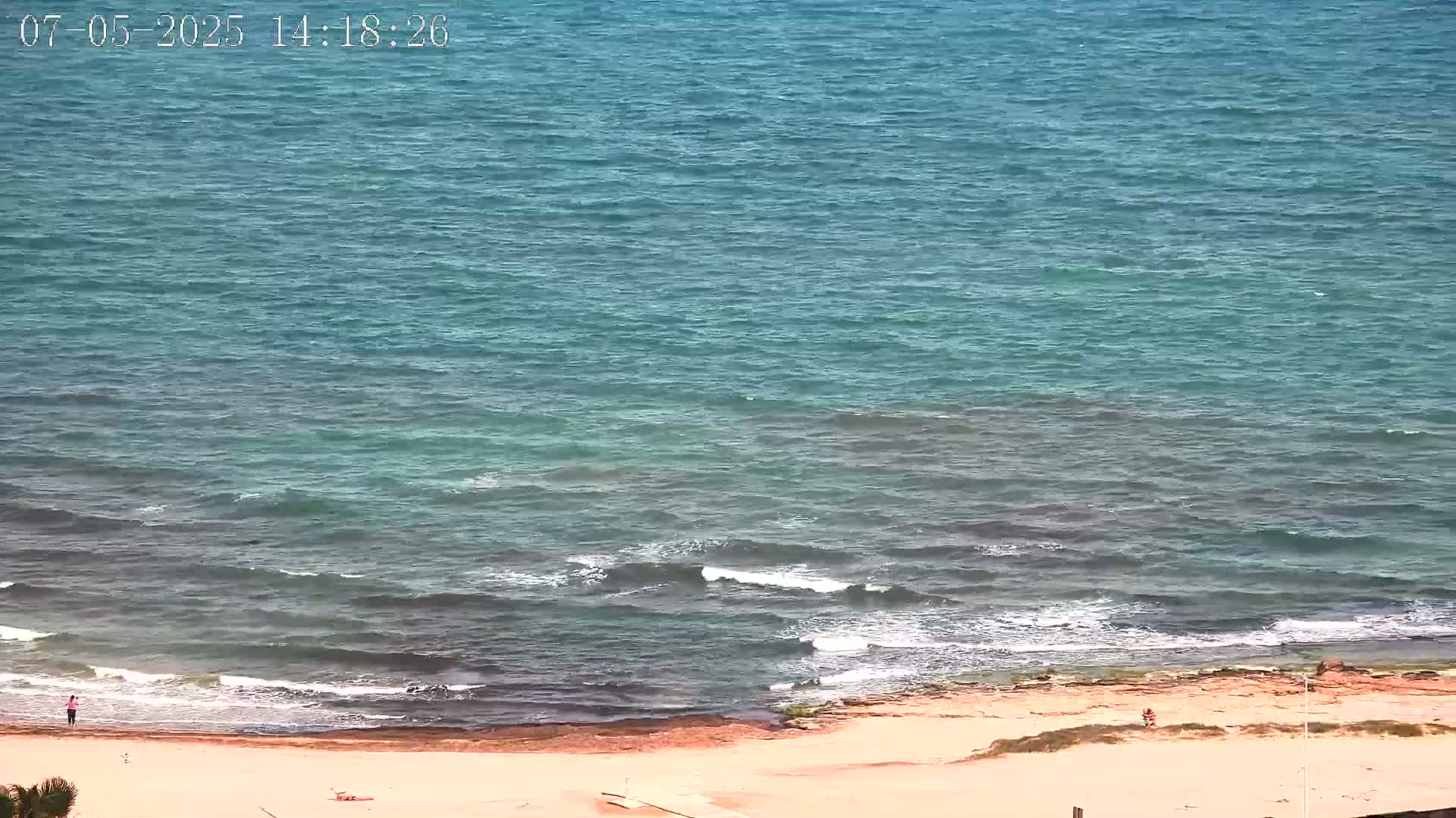 A mostly sunny day at a beach shows a calm sea with small waves lapping a sandy shore where a few people are visible.
