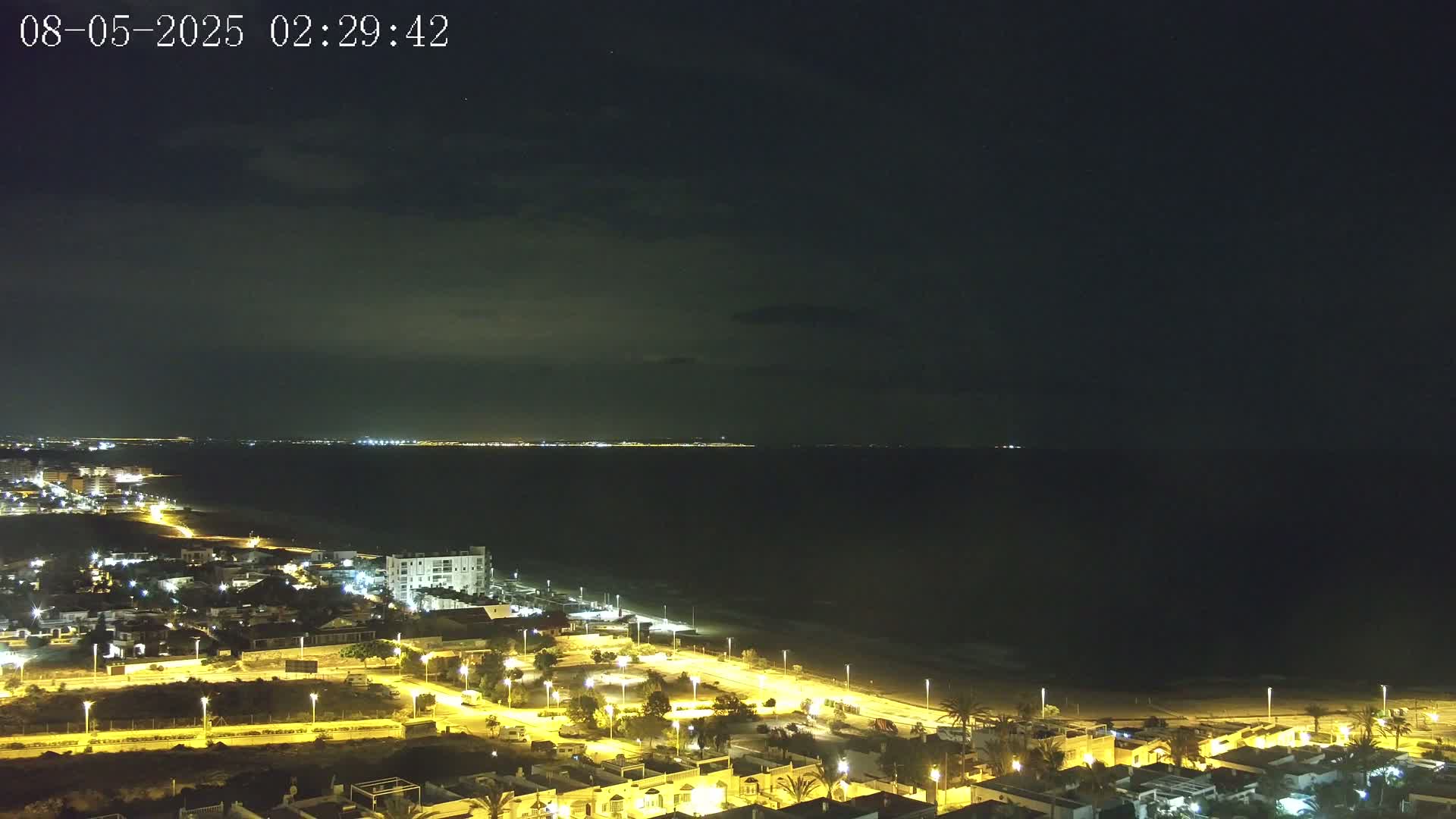 A nighttime coastal scene shows a dark ocean under a mostly clear sky, with a brightly lit town extending along a sandy beach.
