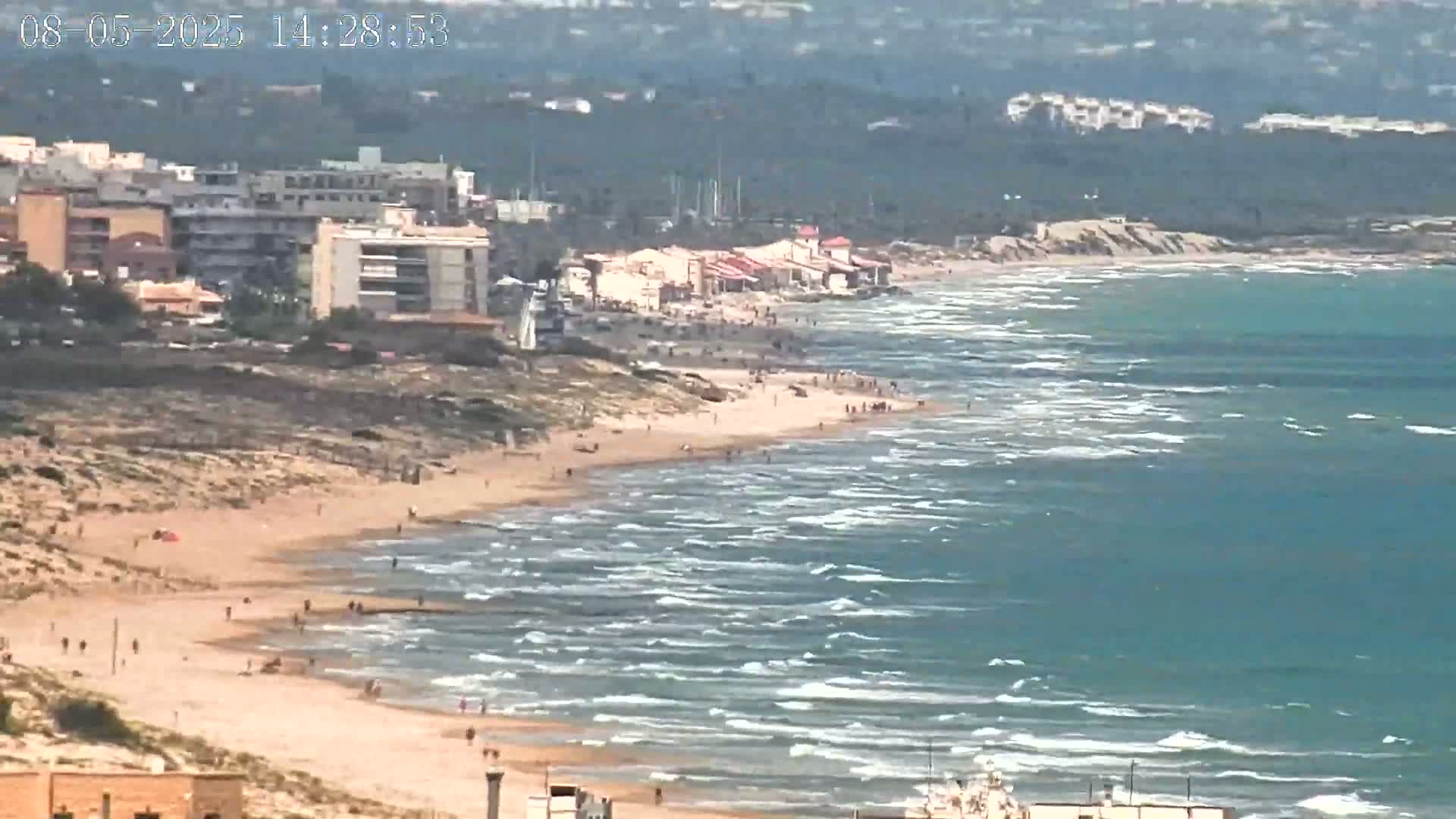 A sandy beach with several people on it stretches along a teal-colored ocean with small waves under a sunny sky, backed by buildings and vegetation.