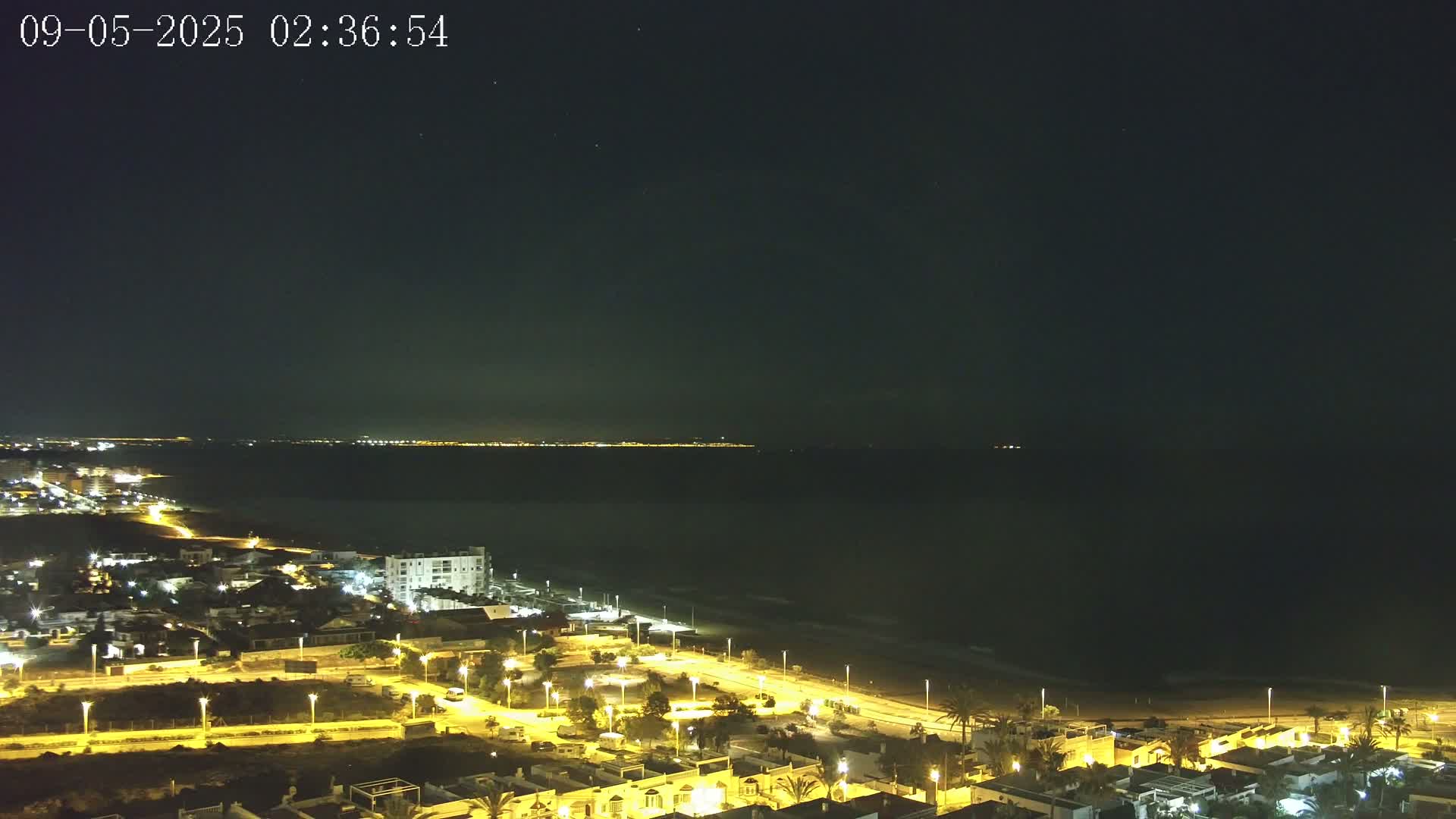 A nighttime coastal town illuminated by streetlights, with a dark ocean and a faintly lit distant shoreline under a clear, starry sky.