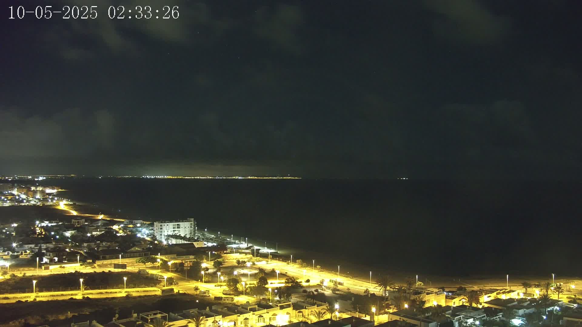 A nighttime coastal town with illuminated streets and buildings overlooks a dark body of water under a mostly cloudy sky.