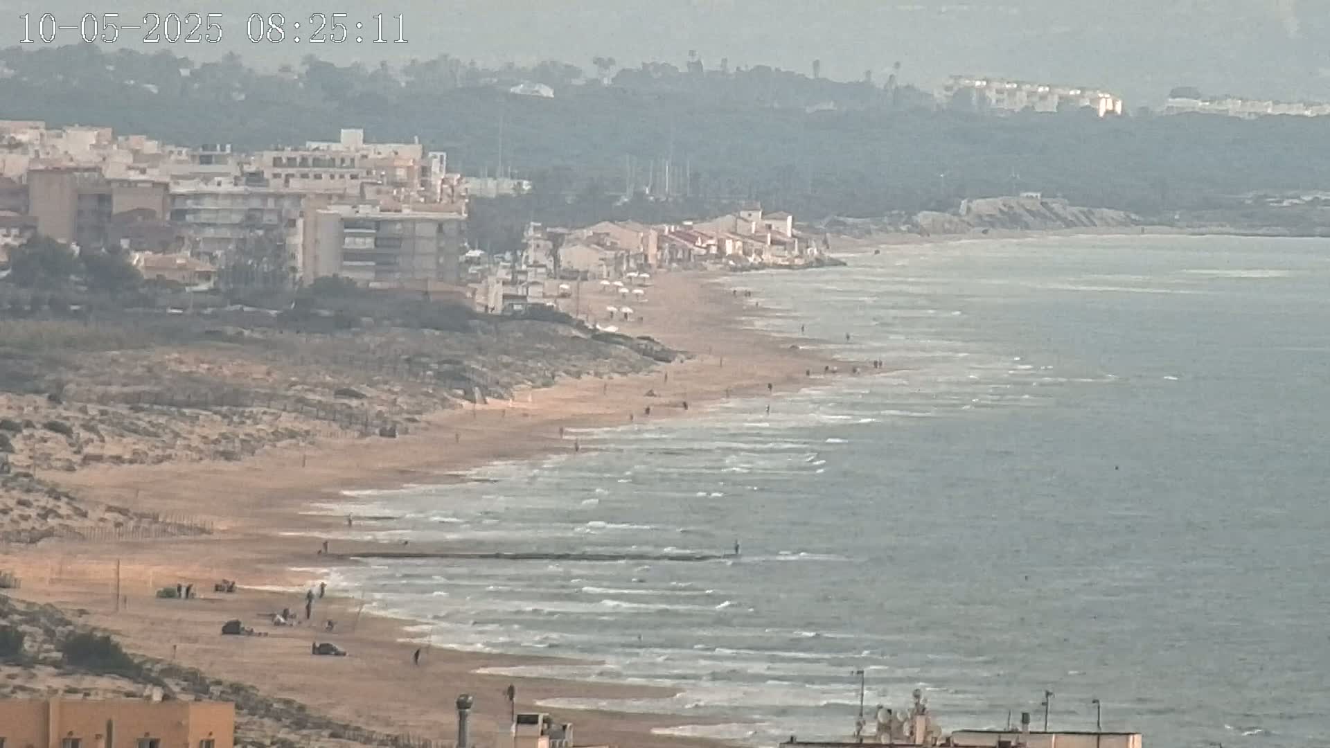 A long sandy beach with a few people on it stretches out to meet a calm, gray sea under an overcast sky, backed by a line of buildings and a hazy hill.