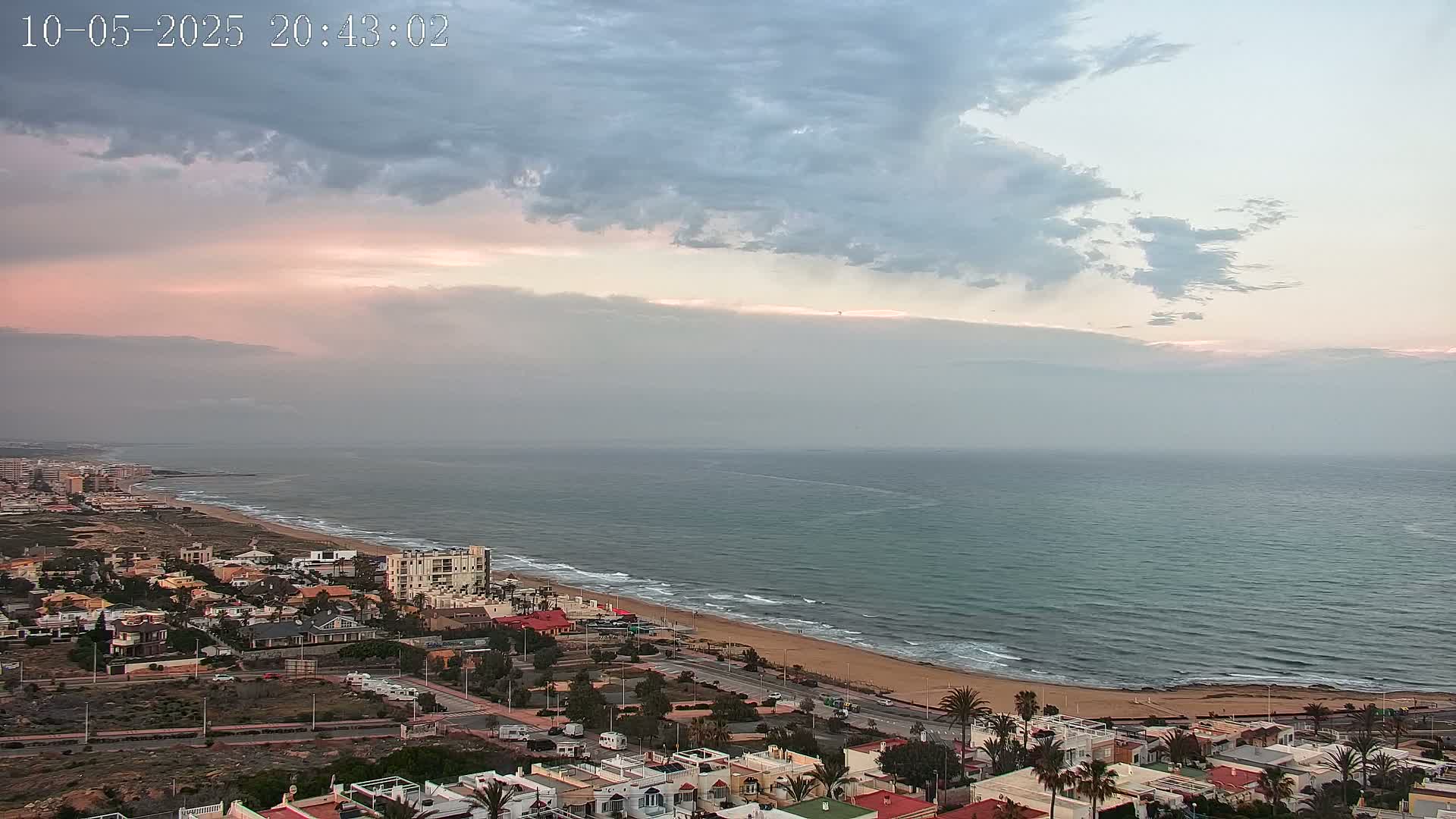 A coastal town with low-rise buildings sits beside a sandy beach and a calm, somewhat hazy ocean under a partly cloudy sky with a hint of pink in the clouds near the horizon.