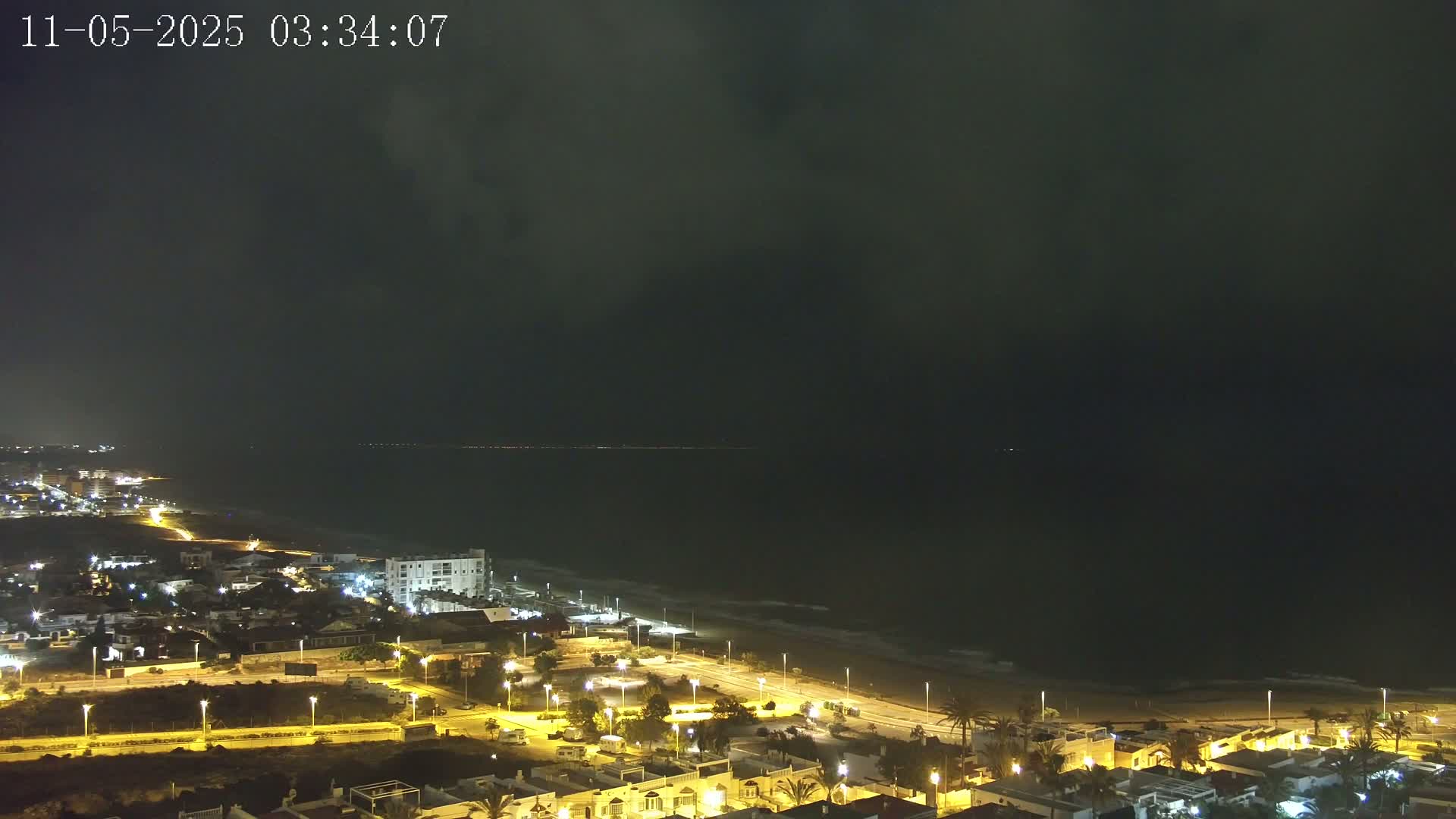 A nighttime coastal town is illuminated, with a dark, cloudy sky above and gently waving water along the beach.