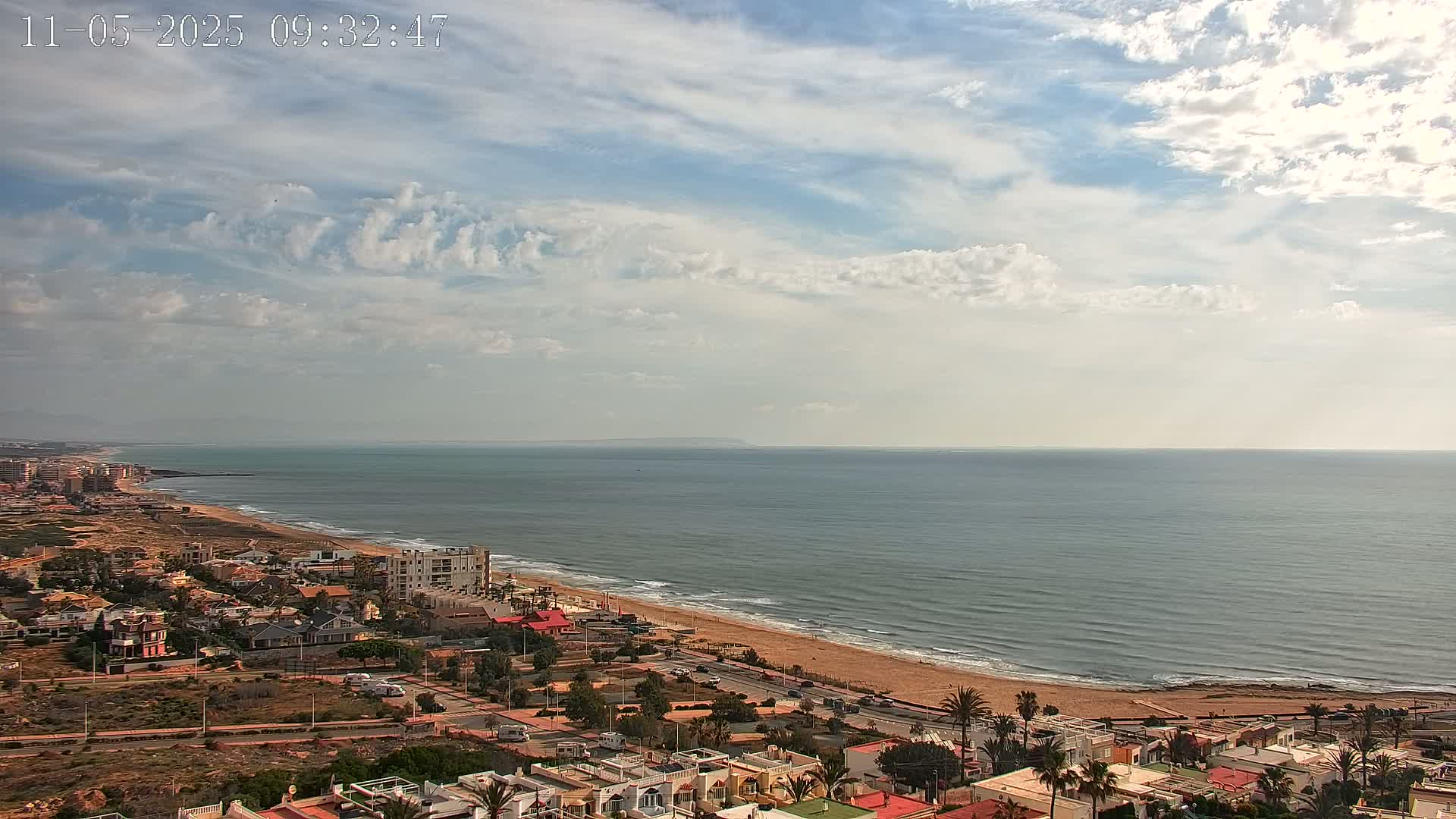 An aerial view shows a calm ocean with a sandy beach and a coastal town under a partly cloudy sky.