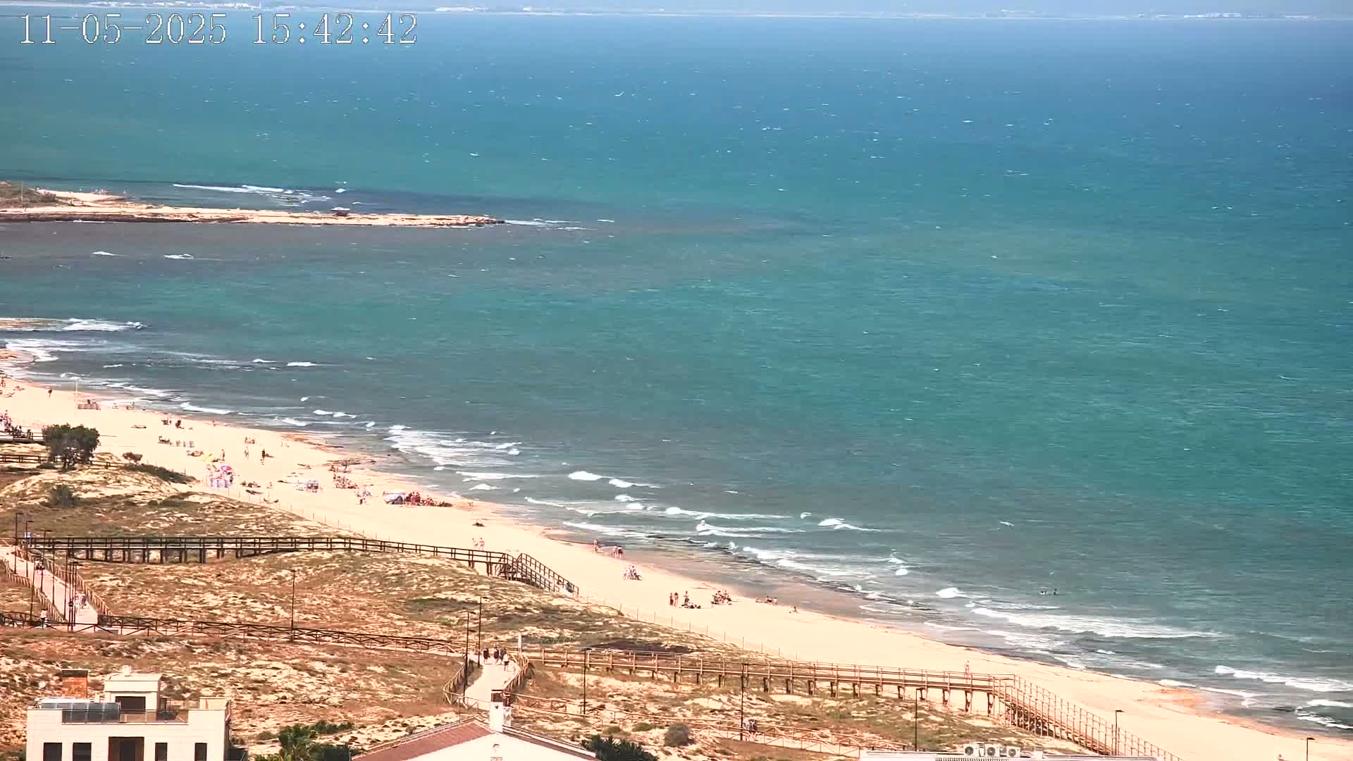 A sandy beach with a few people on it slopes down to a teal ocean with small waves under a sunny sky.
