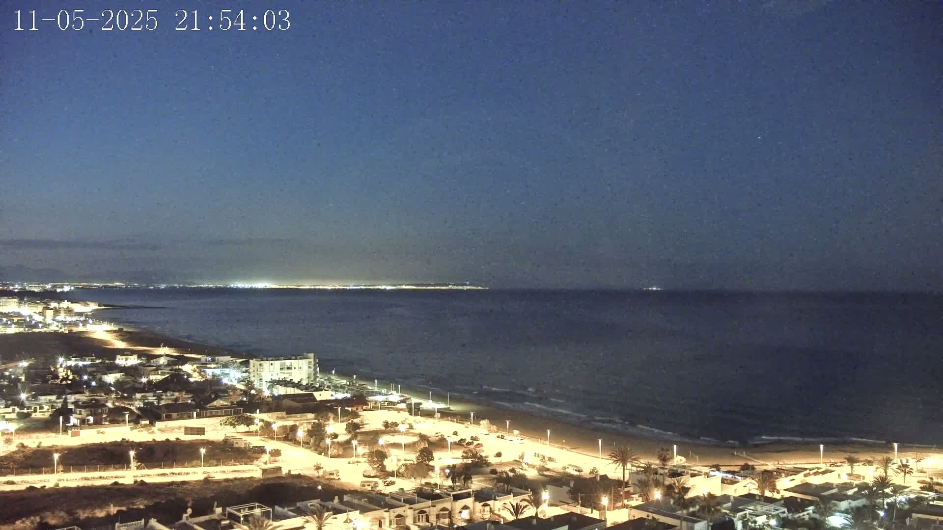 A night-time, coastal view shows a dark blue sea meeting a sandy beach, with a town illuminated by streetlights extending from the beach towards the foreground under a clear, dark sky.