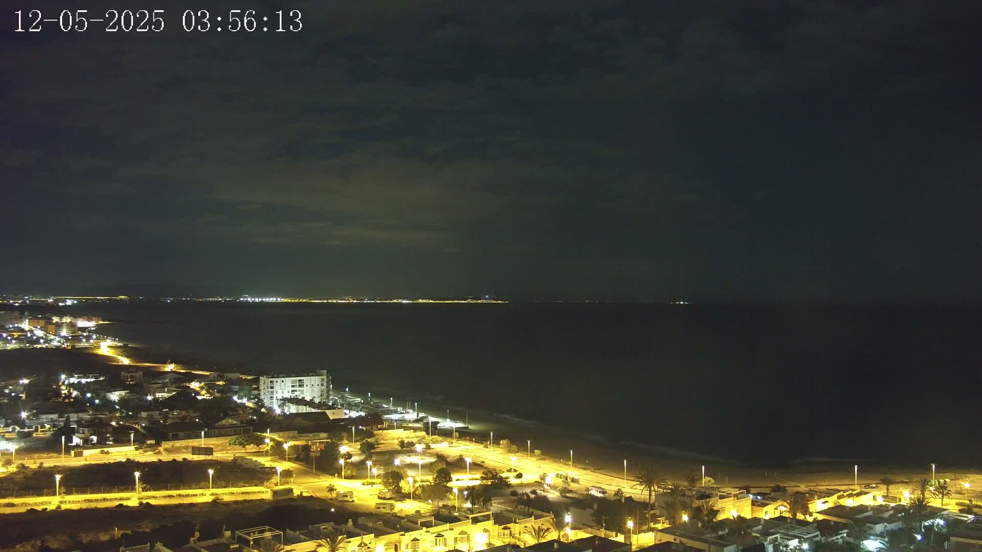 A coastal town at night under a mostly cloudy sky, illuminated by streetlights, overlooks a dark ocean with a distant city skyline faintly visible on the horizon.