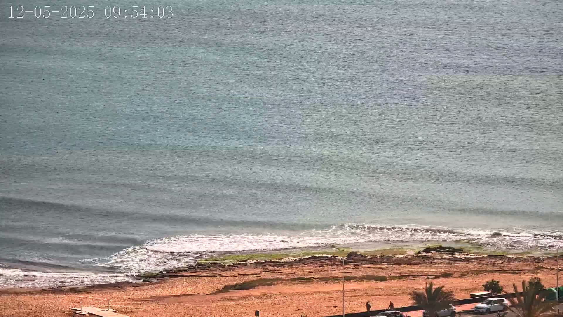 A sandy beach with a rocky shoreline meets a calm, light-blue sea with small waves.