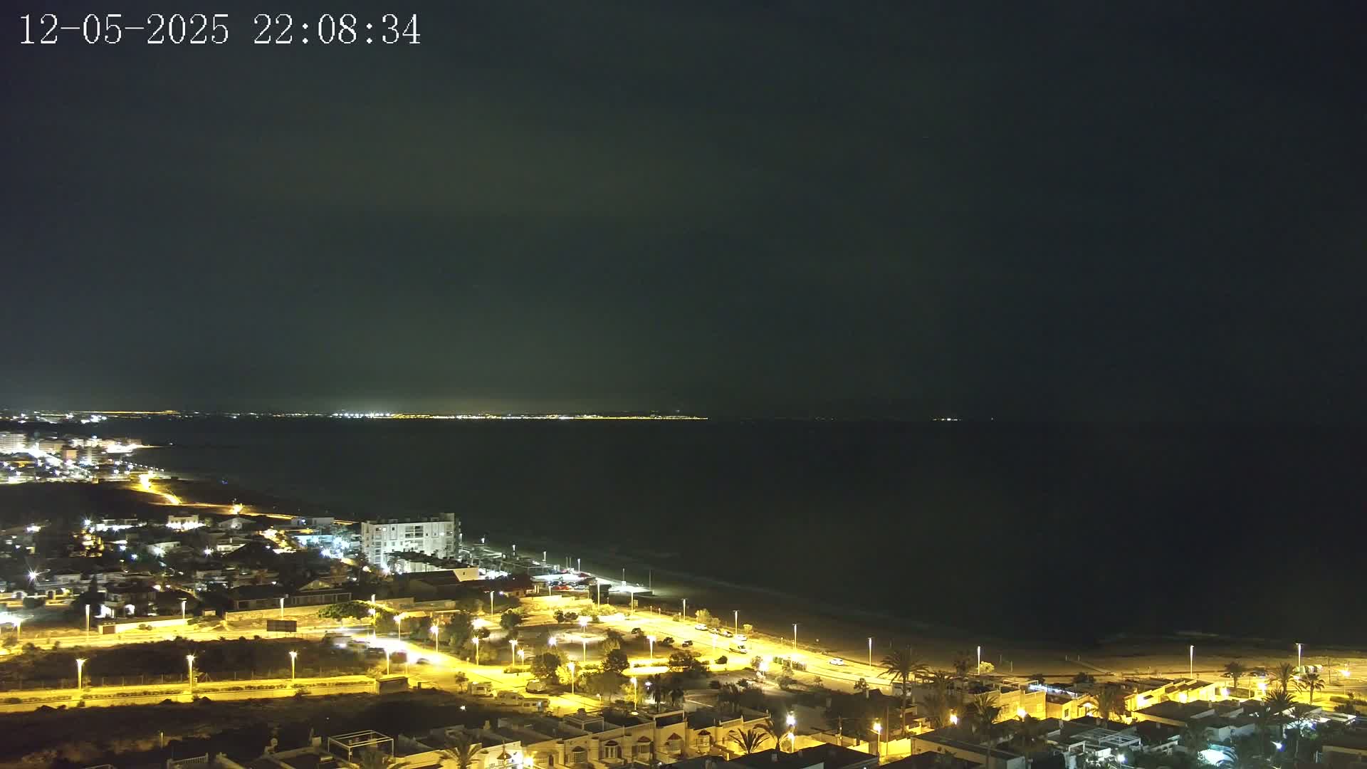 A nighttime coastal town is illuminated, with a dark sea extending to a dimly lit horizon under a clear, dark sky.